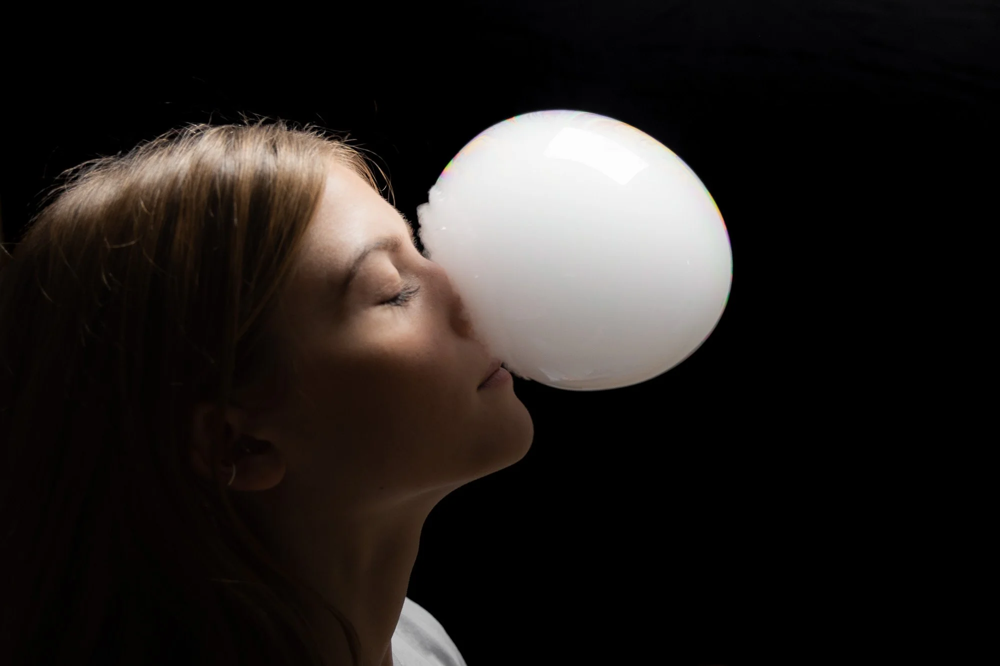 Frau bläst eine weiße Seifenblase mit geschlossenen Augen in dunklem Hintergrund.