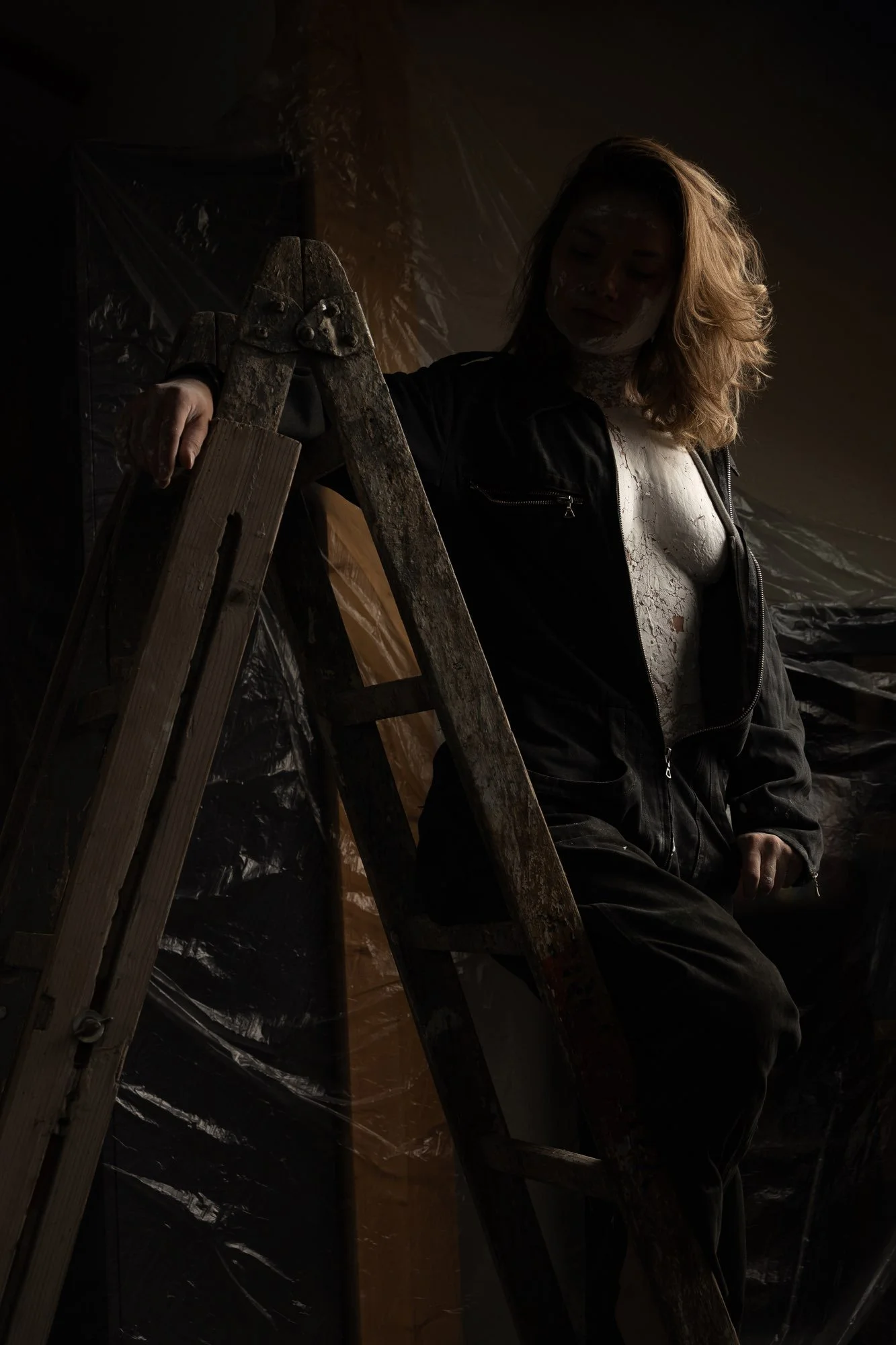 Eine Frau im Schatten posiert neben einer alten, holzfarbenen Leiter in einem dunklen Raum. Sie trägt ein schwarzes Jacke und ein weißes Oberteil.