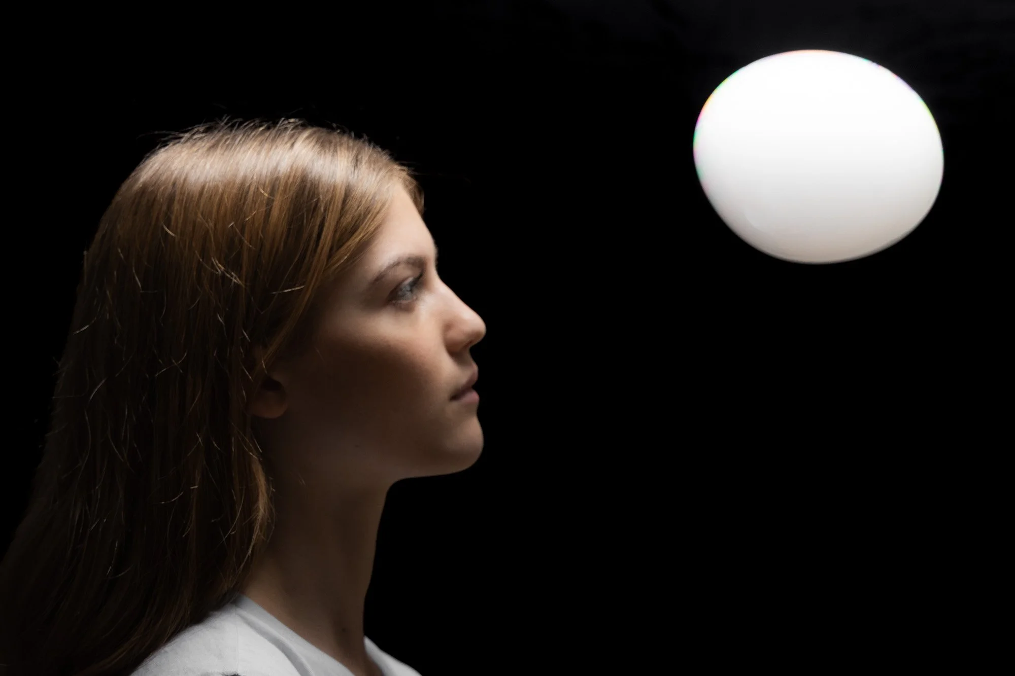 Seitenansicht eines jungen Mädchens mit langen braunen Haaren und weißem Hemd blickt auf einen leuchtenden weißen Ball in dunklem Hintergrund.