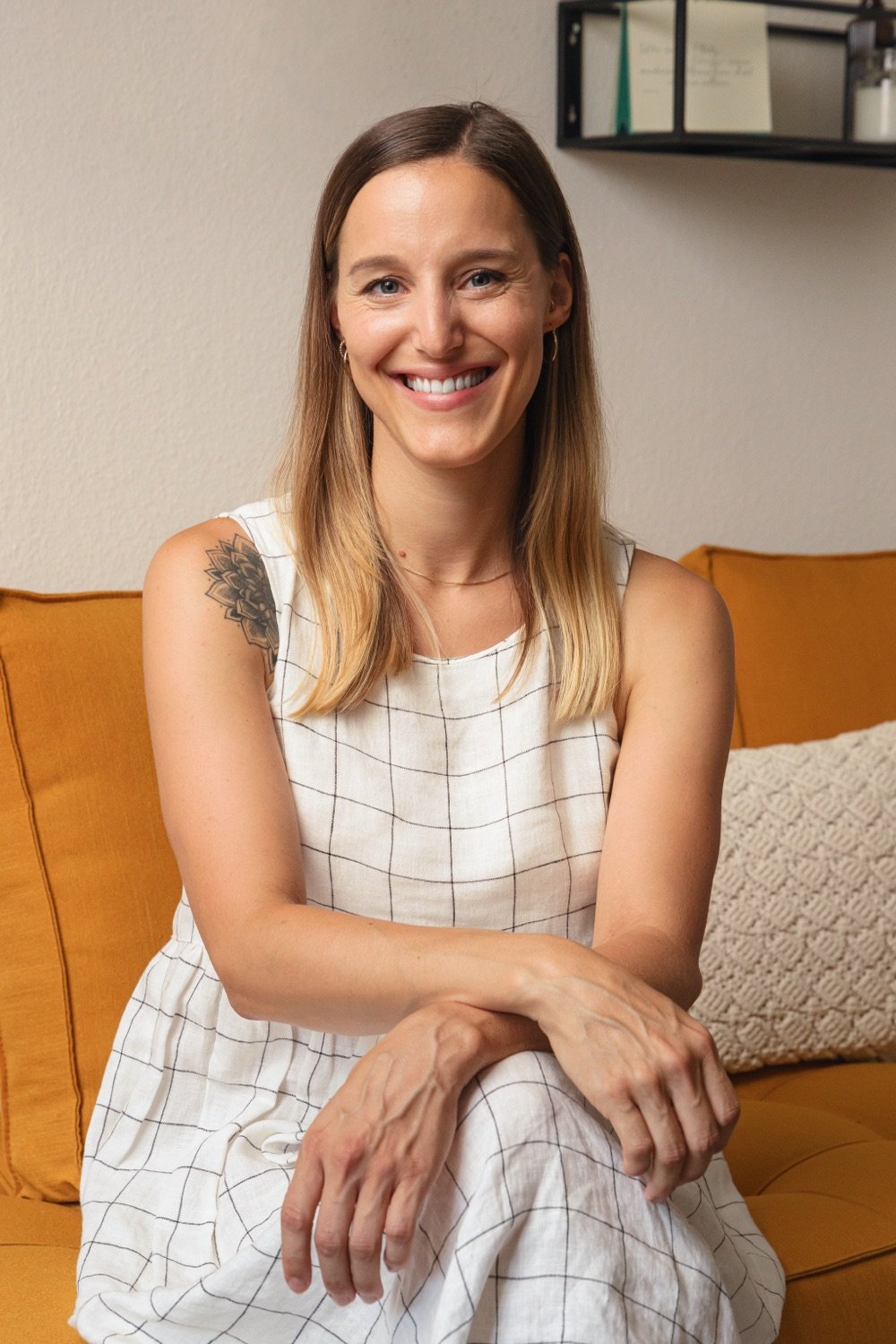 Junge Frau mit lichter Haar, tätowiertem Schulter und freundlichem Lächeln sitzt auf einem orangefarbenen Sofa in einem Wohnzimmer, trägt ein weißes Kleid mit Karomuster.