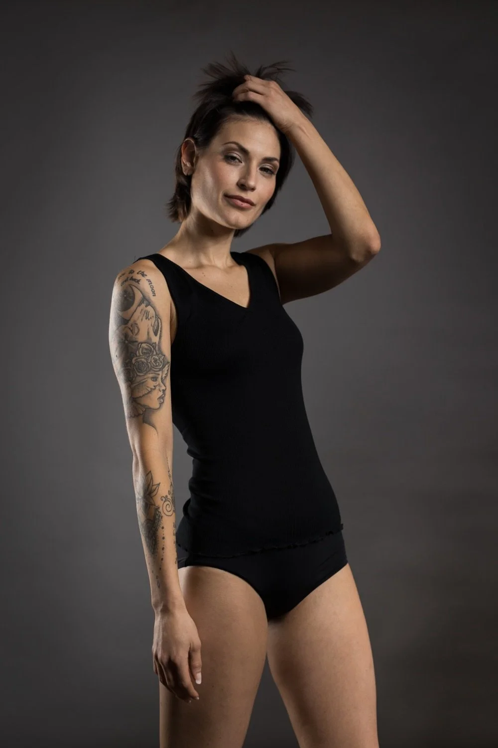 Eine Frauenportraitaufnahme einer Frau in schwarzer Kleidung, mit Tattoos auf dem linken Arm, vor einem dunklen Hintergrund.