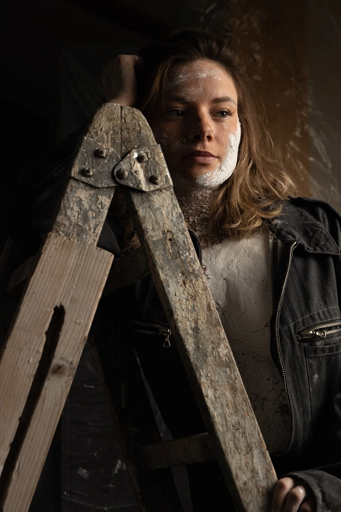 Eine junge Frau steht vor einer dunklen Wand, trägt eine Lederjacke und hat Puder im Gesicht, wirkt nachdenklich, hält eine Leiter in der Hand.