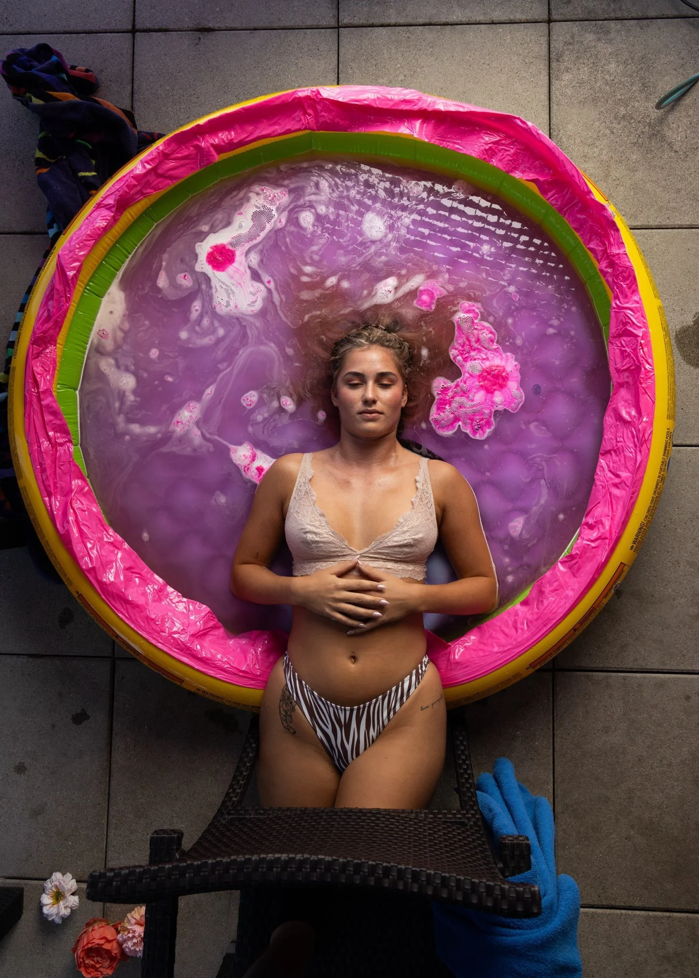 Eine junge Frau liegt entspannt in einem pinkfarbenen aufblasbaren Pool, trägt einem bikini mit Zebramuster, und hat ihre Hände auf dem Bauch. Der Pool befindet sich auf einem grauen Fliesenboden.