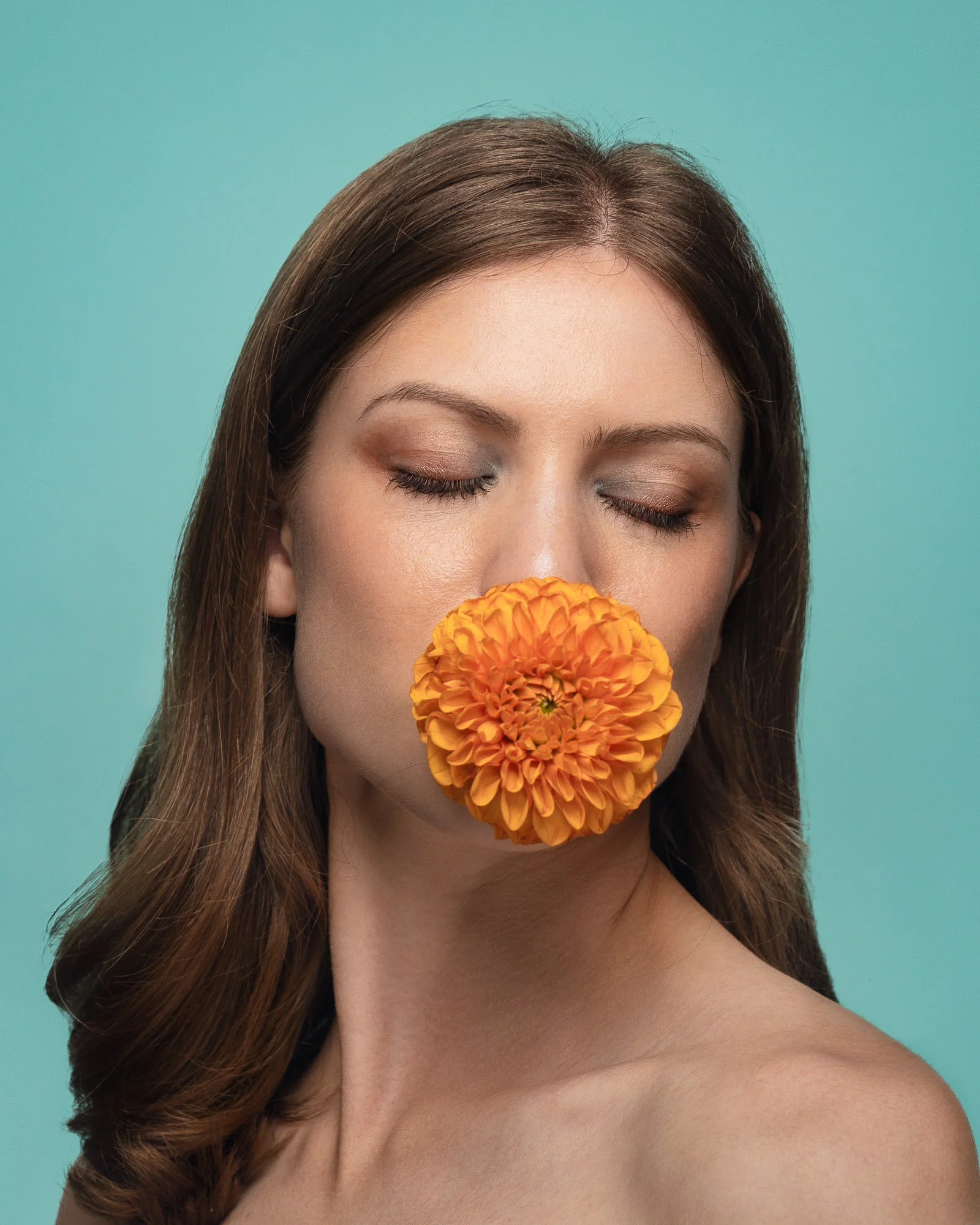 Junge Frau mit geschlossenen Augen und einem orangefarbenen Blumen vor Mund vor einem hellblauen Hintergrund.