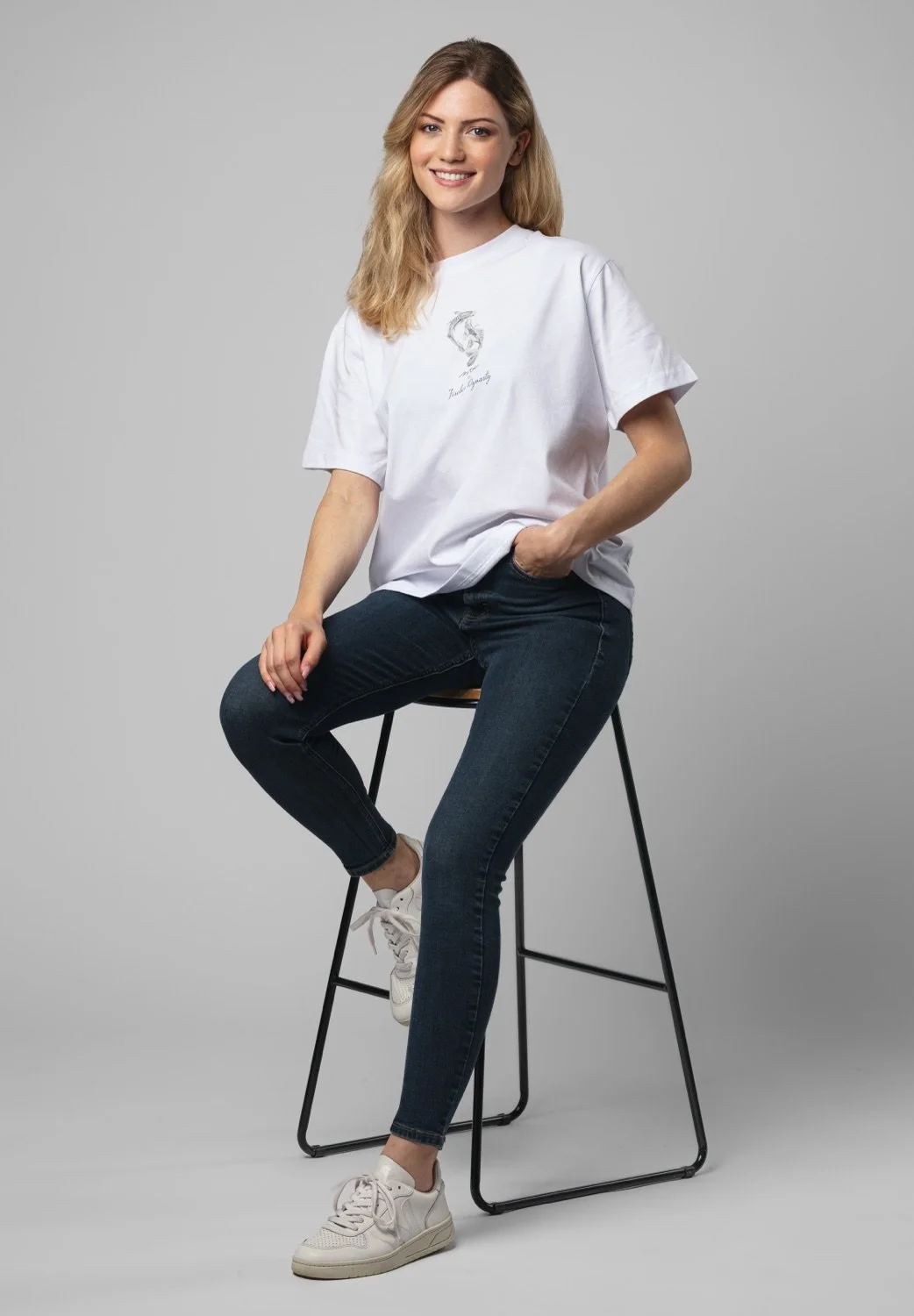 Junge Frau in weißen T-Shirt und blauen Jeans sitzt auf einem Hocker vor grauem Hintergrund.