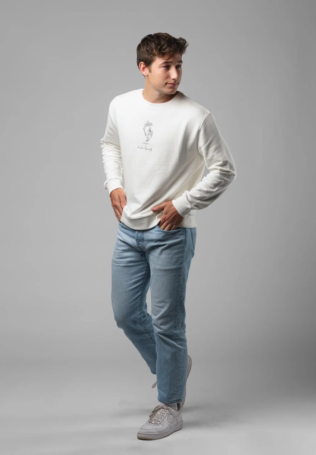 Junge trägt weißen Pullover mit kleiner Grafik und blauen Jeans auf grauem Hintergrund.
