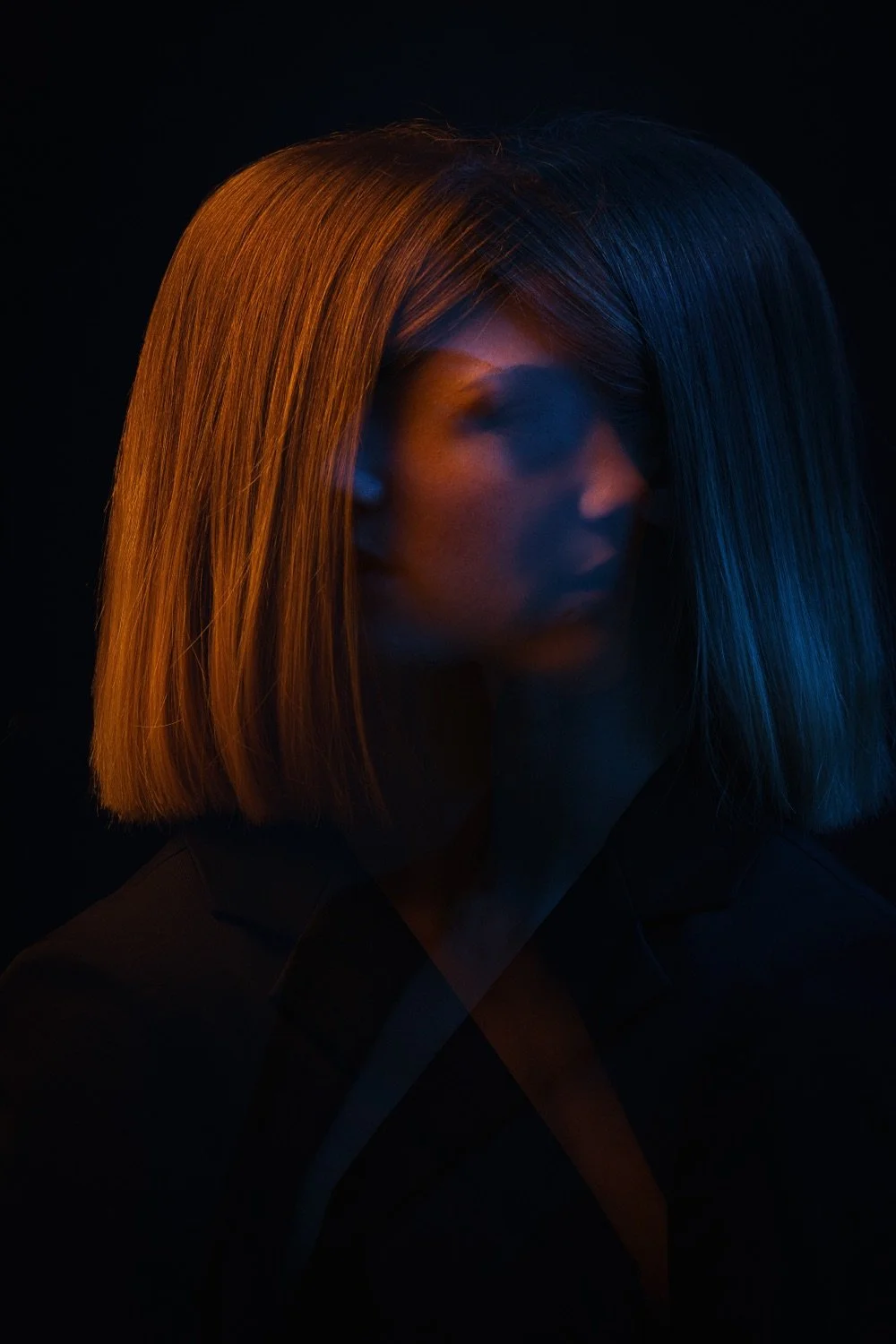 Porträt einer Frau mit geradem, zweifarbig geteiltem Haar in warmem Orange auf der linken und kaltem Blau auf der rechten Seite, bei schwachem Licht im dunklen Hintergrund.