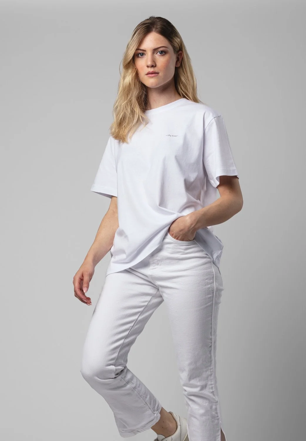 Junge Frau mit blonden Locken trägt ein weißes T-Shirt und weiße Hosen, posiert vor neutralem Hintergrund.