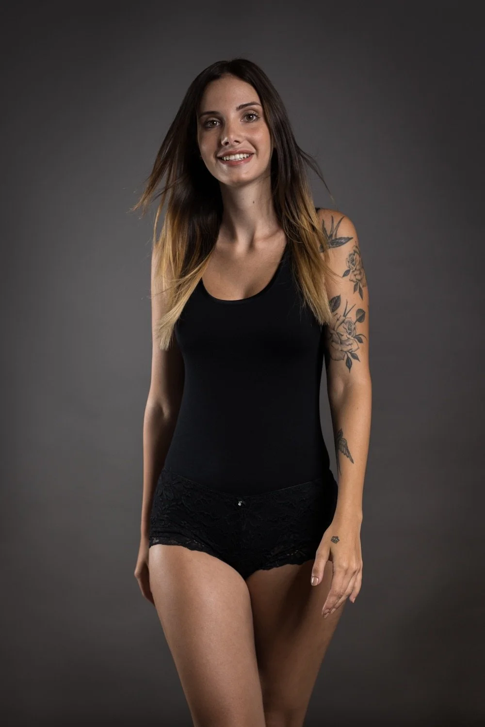 Junge Frau mit Tattoos in schwarzem Tanktop und Shorts vor dunklem Hintergrund.