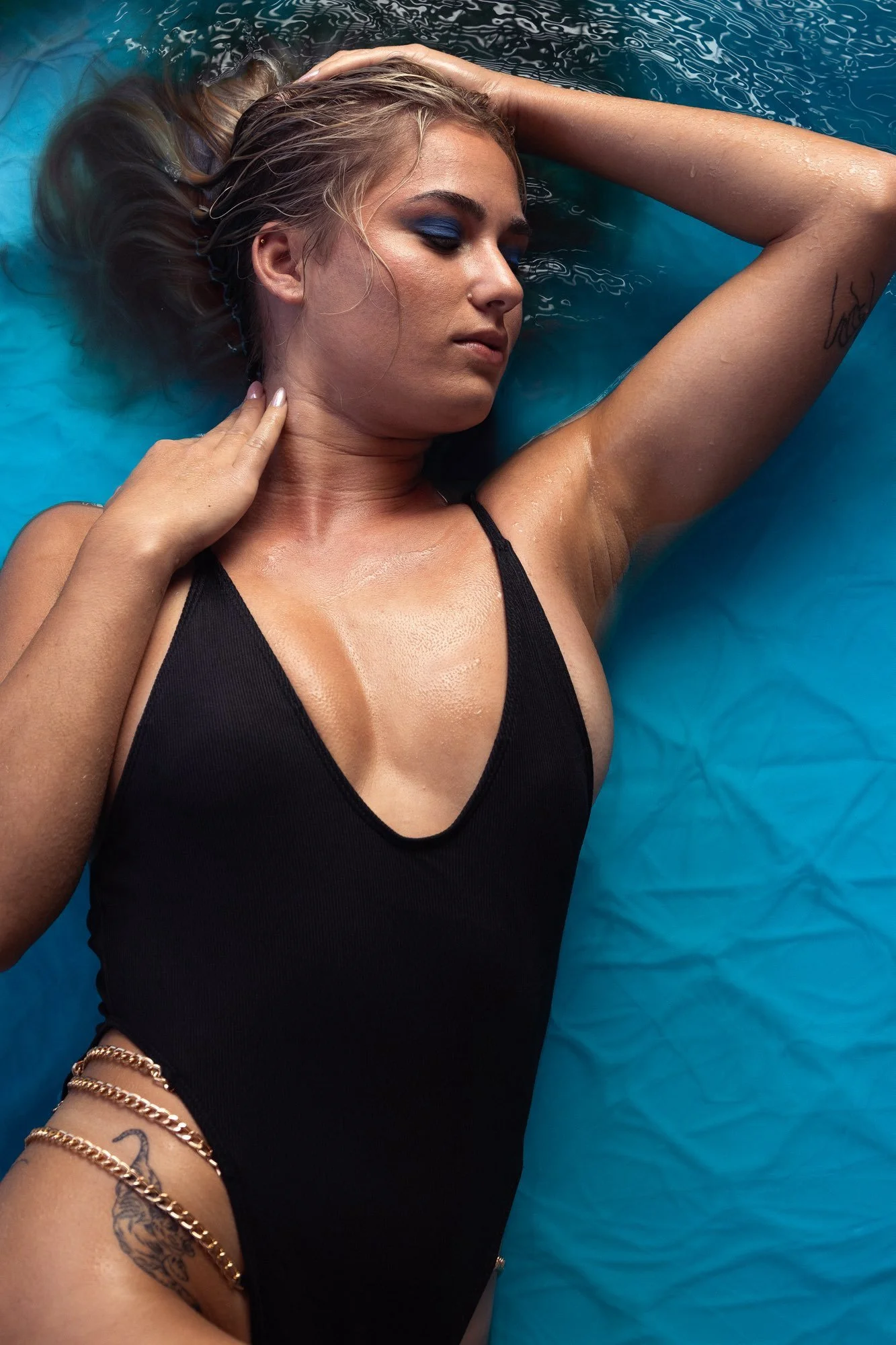 Eine junge Frau liegt entspannt im Wasser, trägt einen schwarzen Badeanzug, hat langes Haar und Tattoos am Arm und Hüfte, mit geschlossenen Augen und einem ruhigen Gesicht.