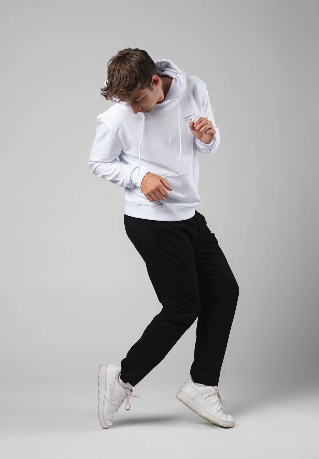 Junge Person tanzt vor hellem grauem Hintergrund, trägt weiße Kapuzenjacke, schwarze Hosen und weiße Turnschuhe.