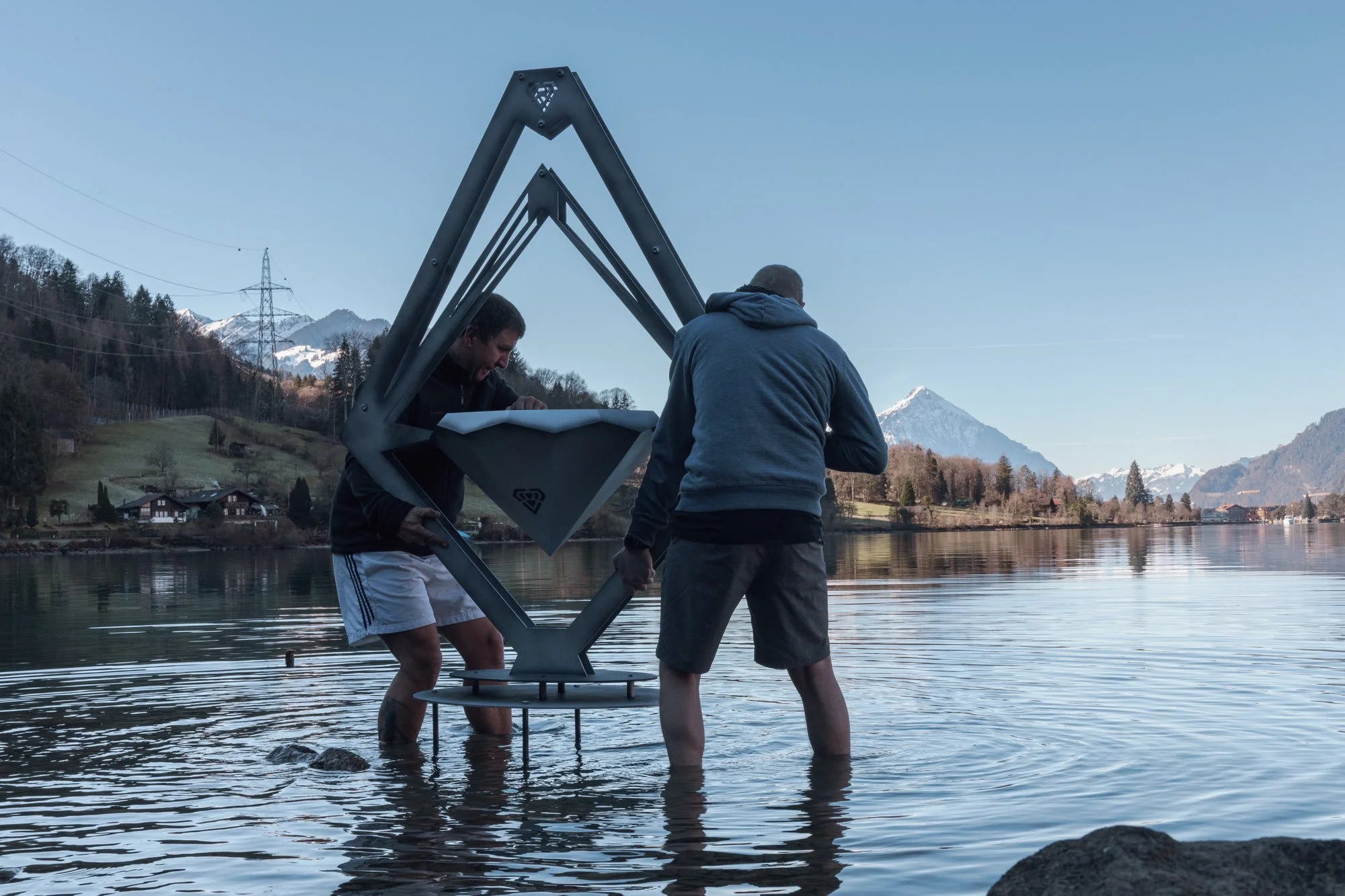 Zwei Männer im Wasser beim Aufbau eines großen Metallrahmens mit einer Box in der Mitte, vor einer Bergkulisse mit Schneegipfeln und einem See.