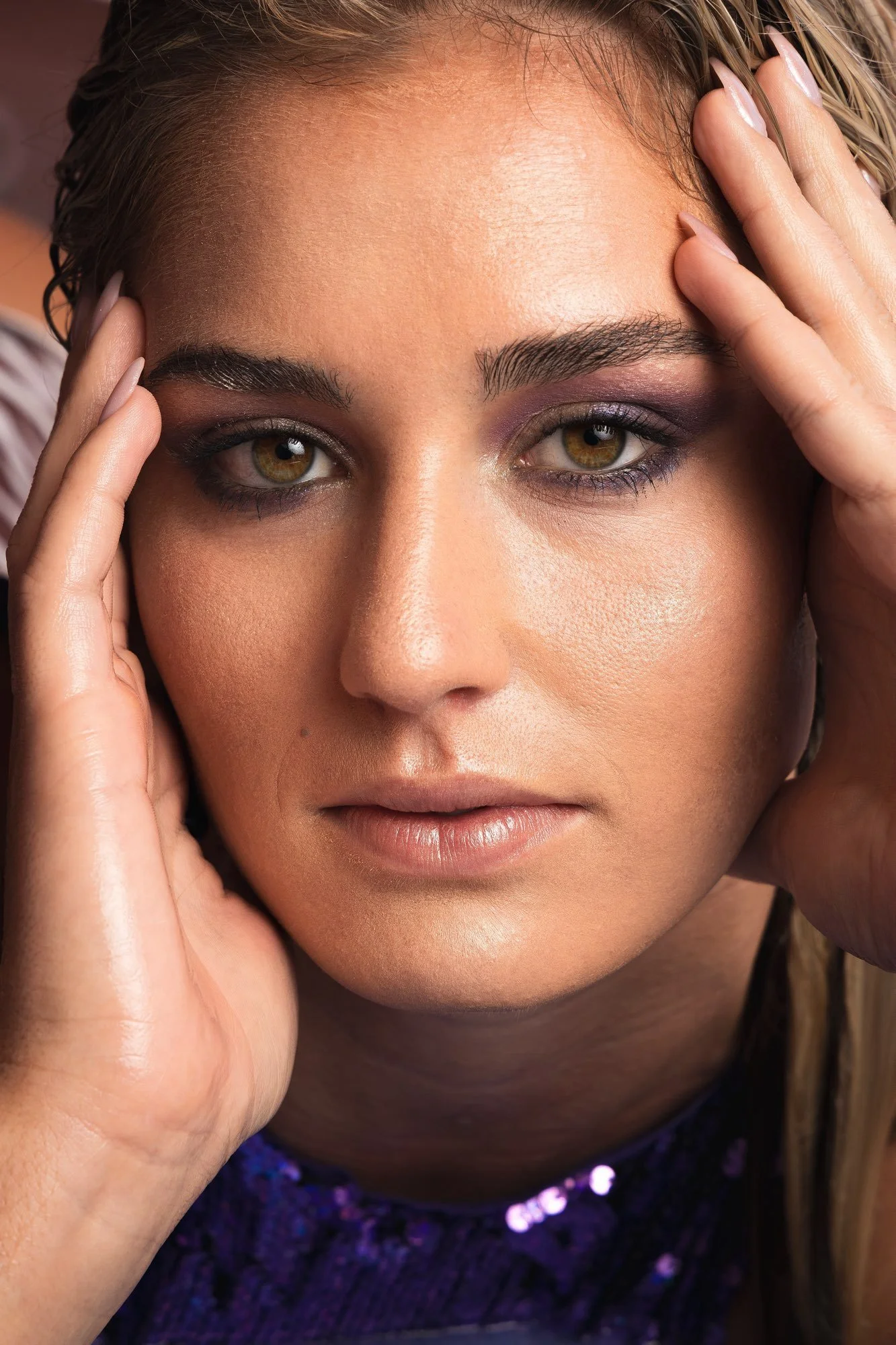 Nahaufnahme eines Frauenporträts mit gepflegter Haut, Make-up und intensiven Blick, die Hände berühren die Kopfhaut, dunkle Kleidung mit Pailletten.