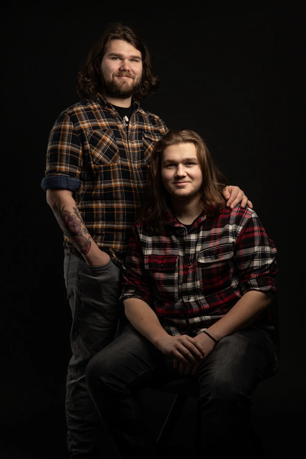 Zwei junge Männer mit langen Haaren, einer sitzt, der andere steht hinter ihm und hat die Hand auf seine Schulter, beide tragen karierte Hemden vor dunklem Hintergrund.