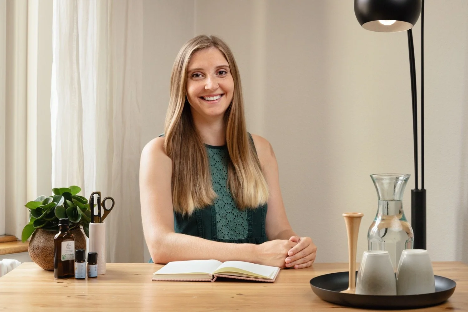 eine junge Frau sitzt an einem Holztisch, lächelnd, mit einem offenen Buch vor ihr, neben ihr sind Pflanzen, kleine Flaschen, und ein schwarzes Tablett mit mehreren Gläsern und Dekorationsgegenständen, im Hintergrund ein Fenster und eine Stehlampe.