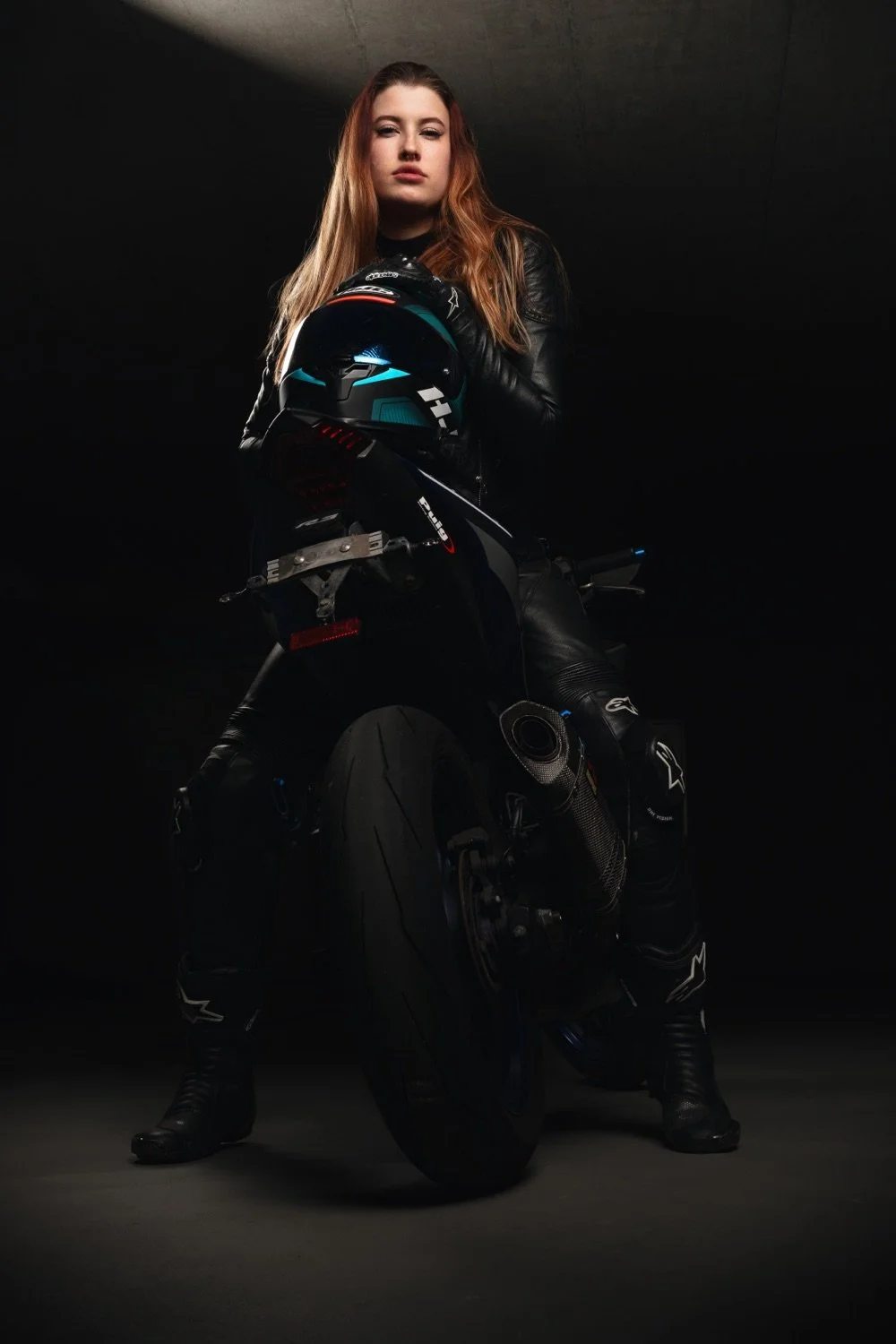Eine junge Frau in schwarzer Motorradbekleidung steht neben einem schwarzen Rennrad, hält einen Rennradhelm in den Armen, liegt in einem dunklen Raum, mit dunklem Hintergrund und subtiler Beleuchtung.