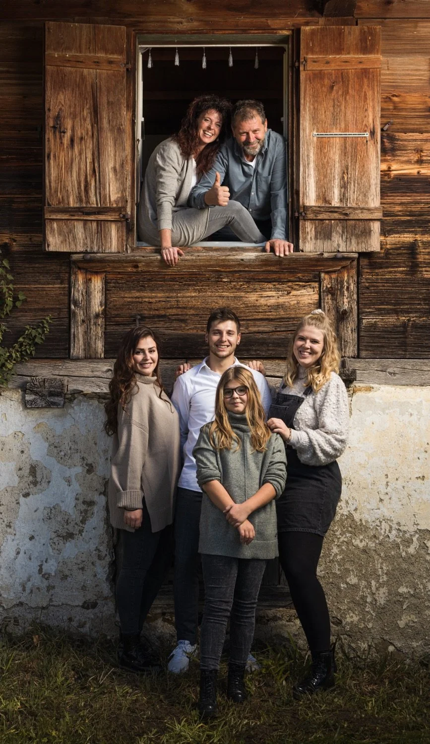 Eine mehrköpfige Familie vor einem alten Holzhaus. Mehrere Familienmitglieder sind im Fenster des Hauses und auf dem Boden darunter, alle lachen und posieren für die Kamera.