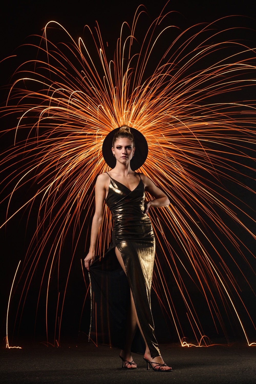 Frau in einem schwarzen, glänzenden Abendkleid mit hoher Slit, trägt einen schwarzen Hut, vor einem Hintergrund mit Feuerwerk.