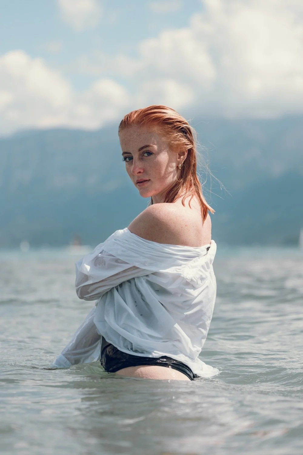 Eine Frau mit roten Haaren trägt ein nasses weißes Hemd und schwarze Bikini-Höschen, steht im Wasser eines Sees oder Meeres, im Hintergrund sind Berge und Wolken am Himmel.