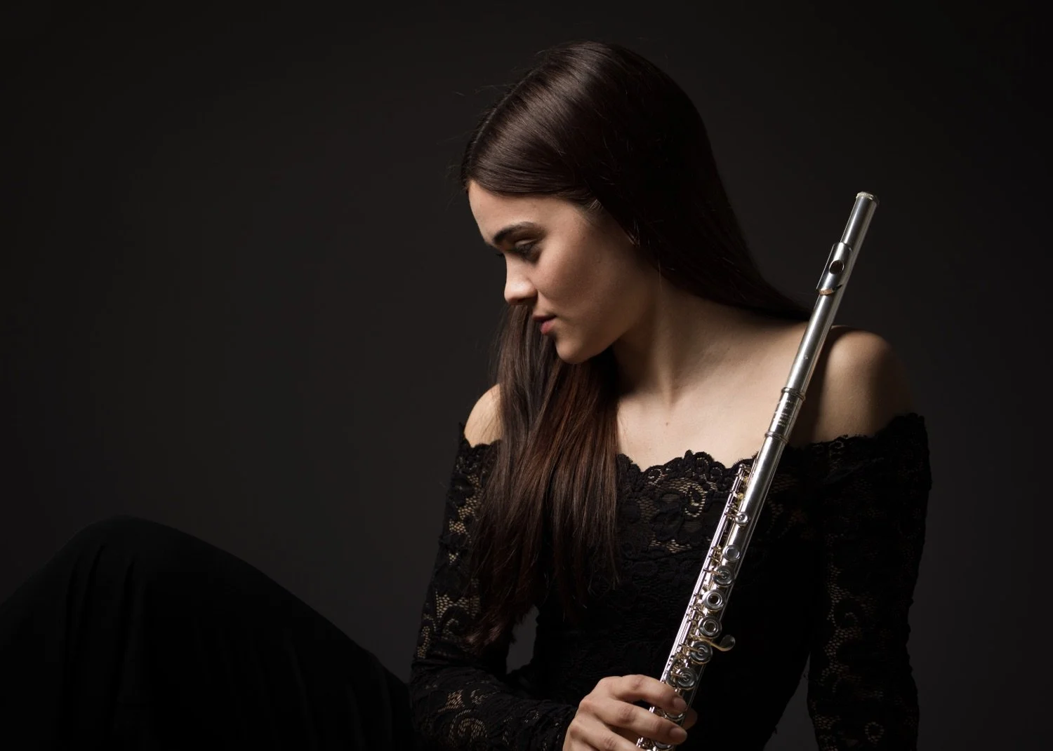 Junge Frau mit dunklen Haaren trägt ein schwarz-elastisches Kleid und hält eine silberne Flöte vor einem schwarzen Hintergrund.