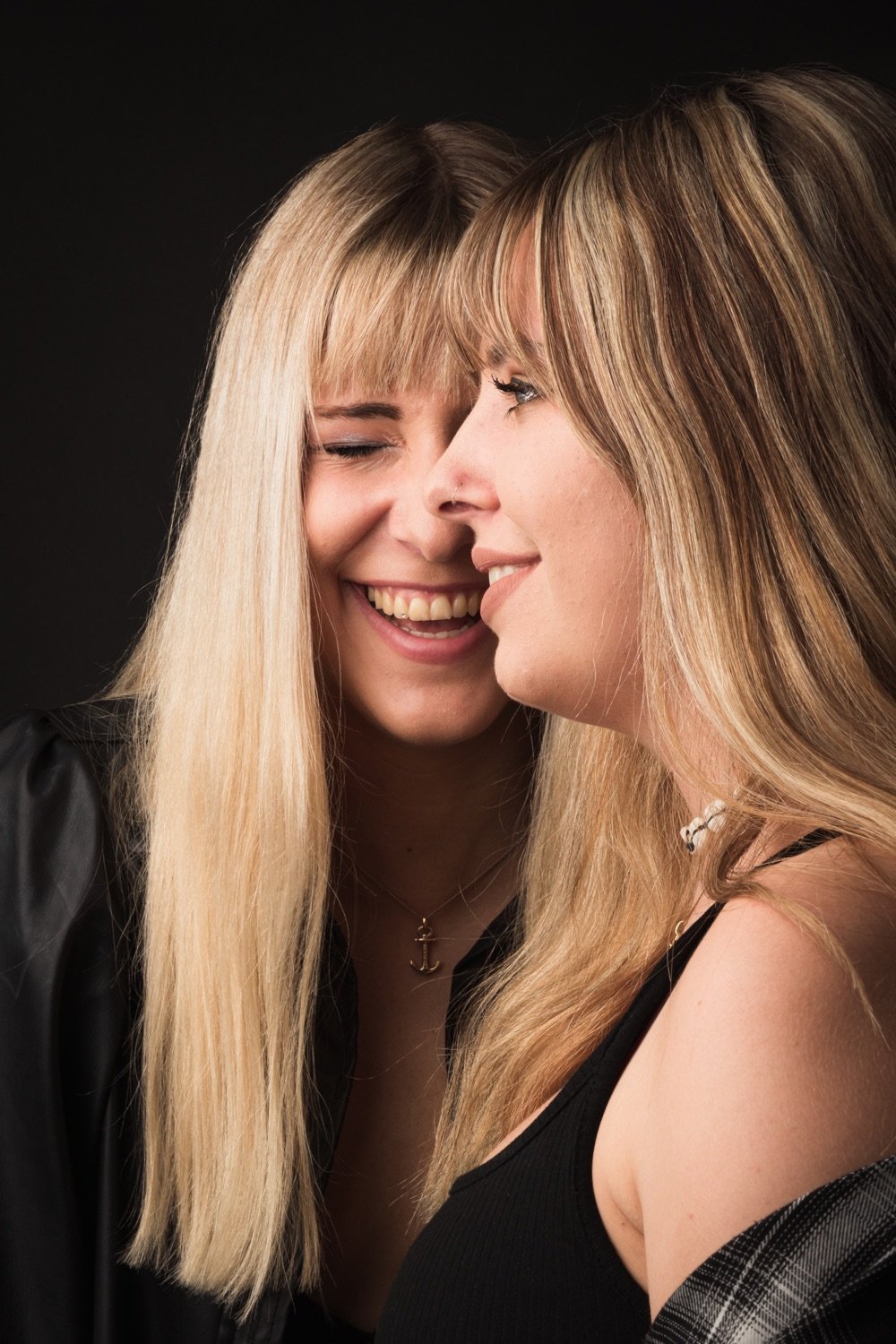 Zwei Frauen lachen und sprechen eng beieinander auf einem dunklen Hintergrund.