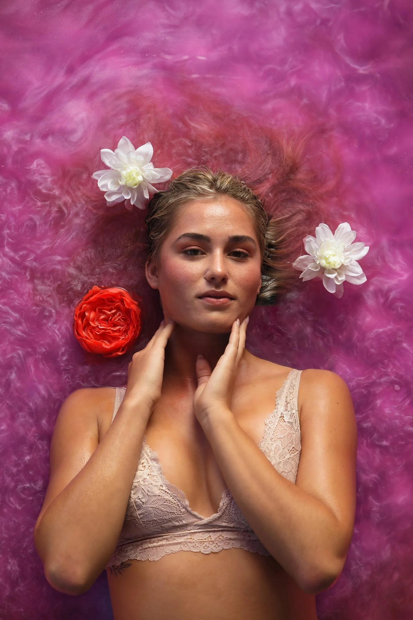 Eine Frau liegt auf einem pinken, watteartigen Untergrund, umgeben von weißen und roten Blumen, mit einer entspannten Gesichtsausdruck und den Händen am Hals.