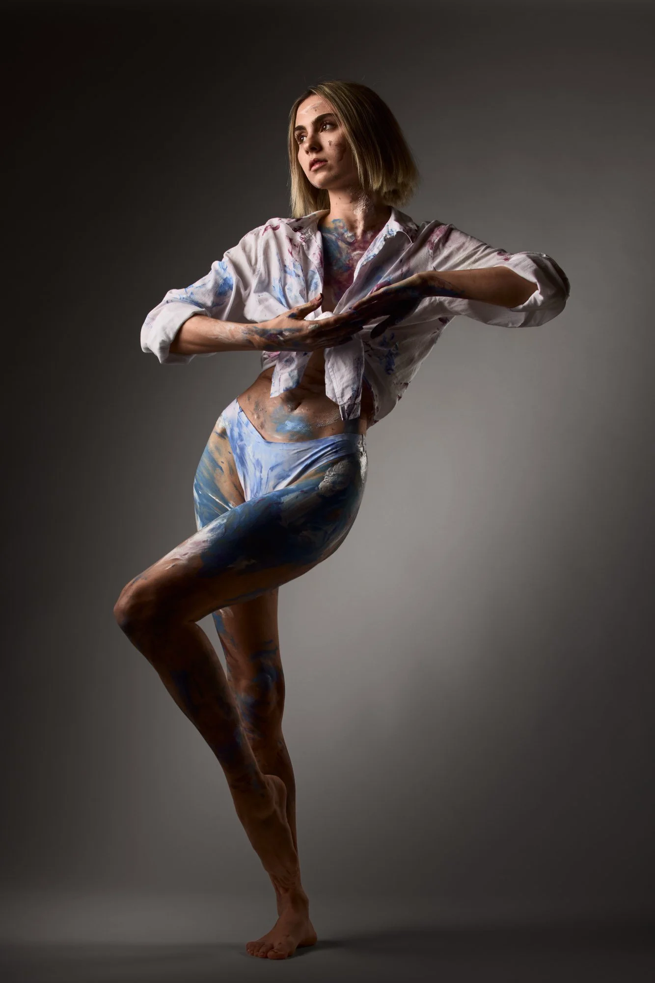 Junge Frau in weißem Hemd, die in Pose steht, mit Körper bemalt in blauen, weißen und braunen Farben, vor grauem Hintergrund.