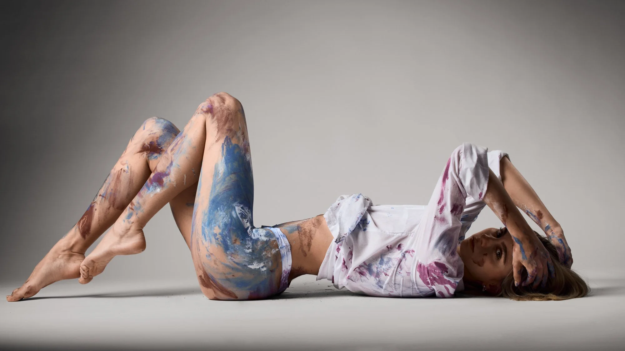 Frau liegt auf dem Boden, bemalt mit Farbklecksen, mit einem weißen Hemd, das ebenfalls Farbflecken hat.