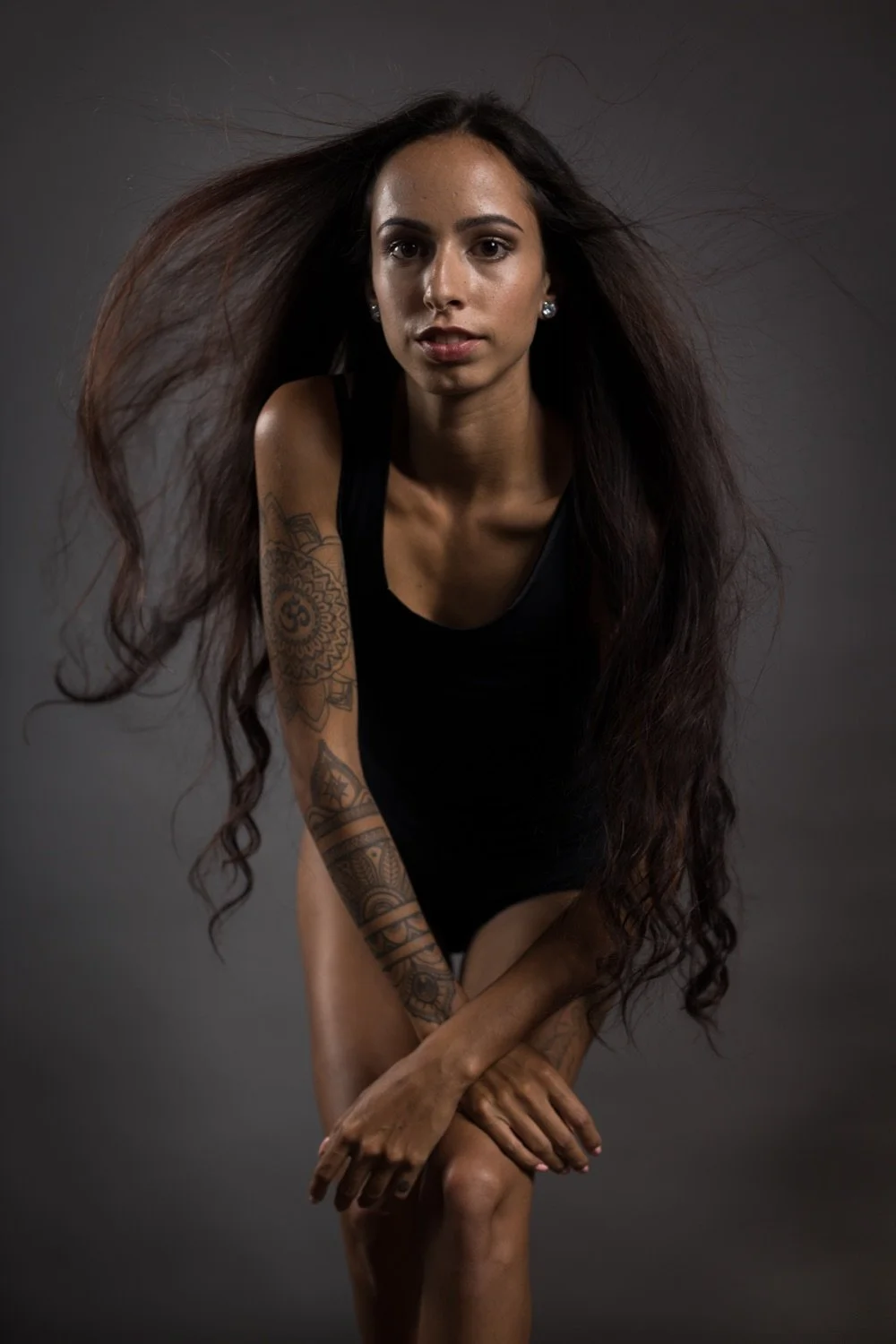 Frau mit langem, lockigem Haar trägt schwarze Kleidung vor grauem Hintergrund.