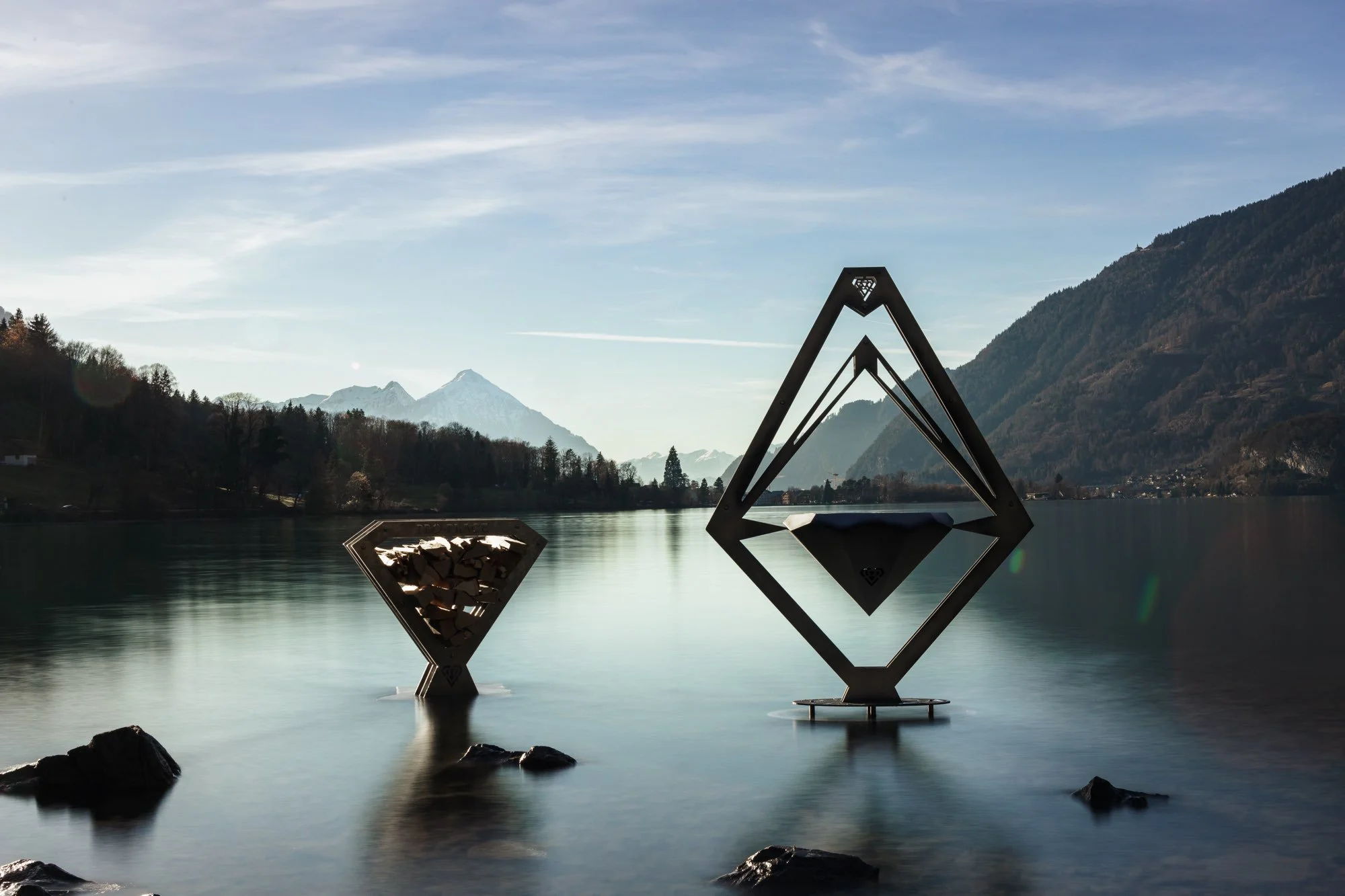 Zwei moderne Skulpturen im Wasser vor einer Bergkulisse mit Schnee, umgeben von Bäumen und Bergen.
