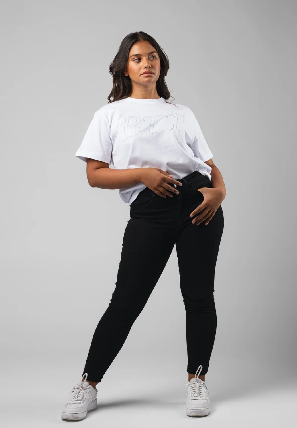 Eine junge Frau in weißen Turnschuhen, schwarzen Hosen und einem weißen T-Shirt steht vor einem neutralen grauen Hintergrund.