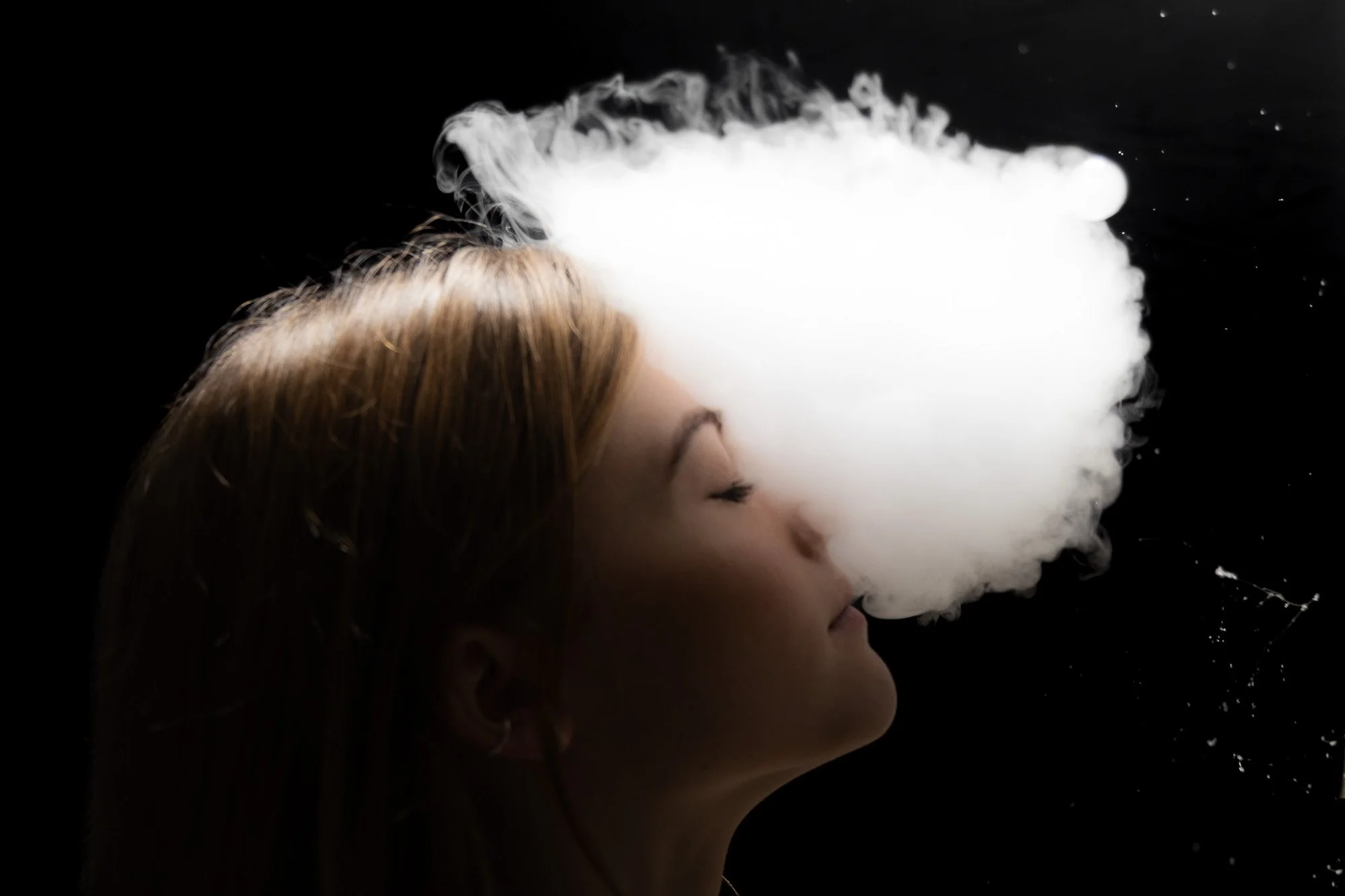 Frau mit geschlossenen Augen, die weißen Dampf aus dem Mund in einer dunklen Umgebung ausstößt.