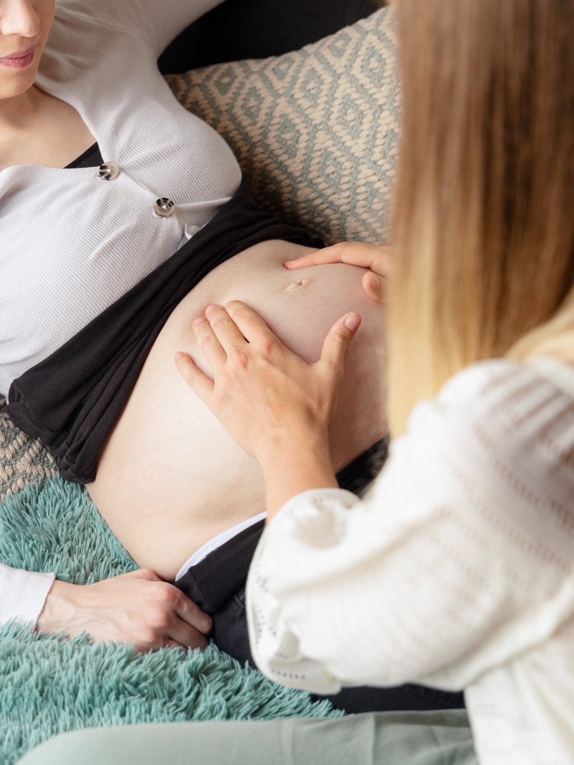 Eine schwangere Frau liegt entspannt auf einer Couch, während eine andere Person, vermutlich eine Hebamme oder Ärztin, den Bauch der schwangeren Frau untersucht. Der Fokus liegt auf dem Bauchbereich.