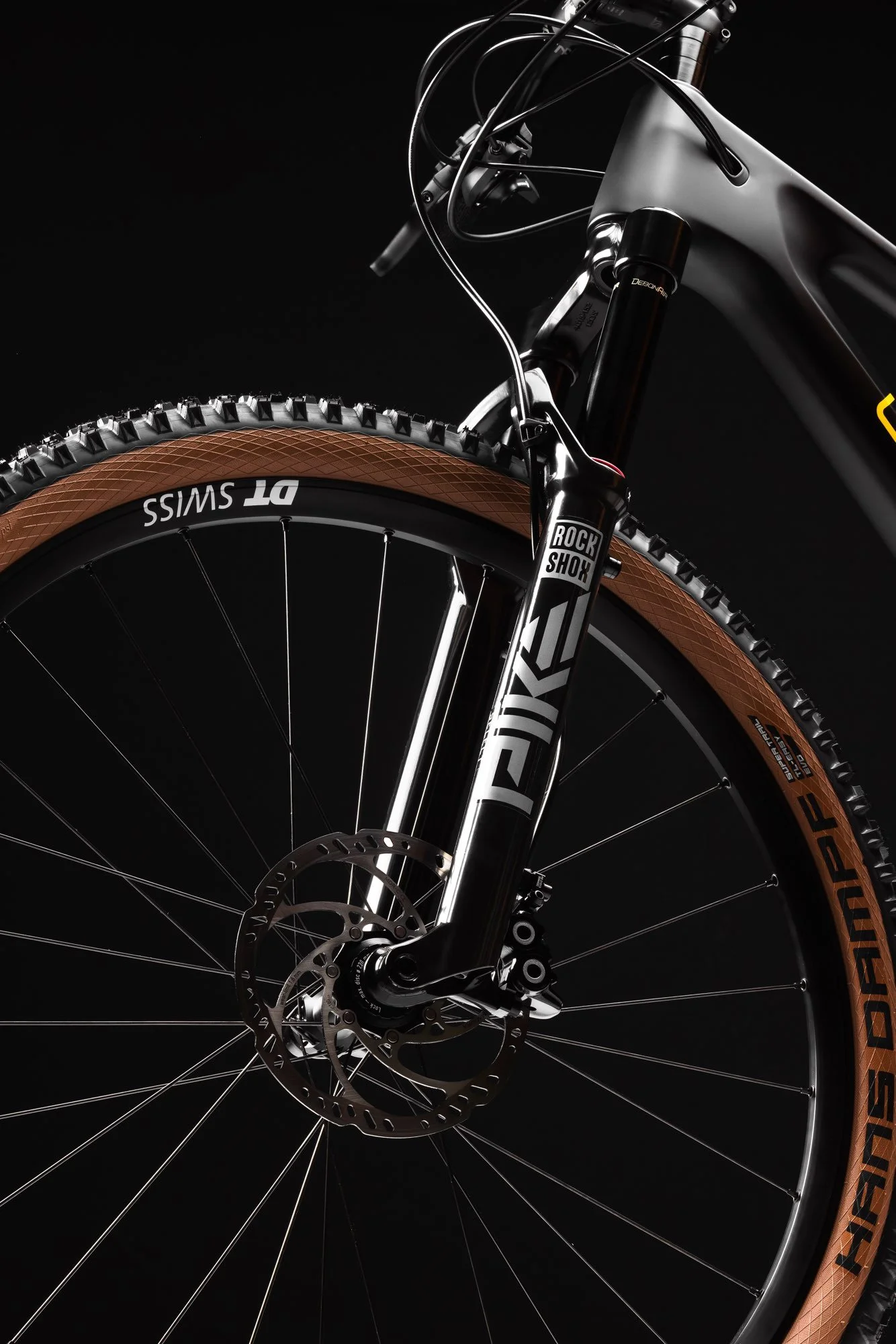 Nahaufnahme des Vorderkotflügels eines Mountainbikes mit schwarzen Rahmen und braunen Reifen. Es zeigt die Gabel, Bremsscheibe und die Kabel. Der Hintergrund ist schwarz.