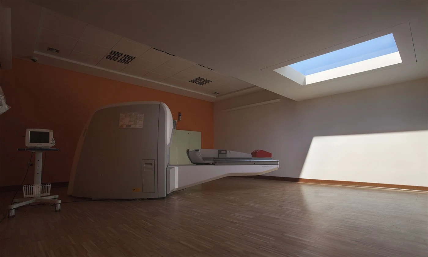 Empty room with a large medical imaging machine near a peach-colored wall, a monitor on a stand, and a skylight allowing natural light.