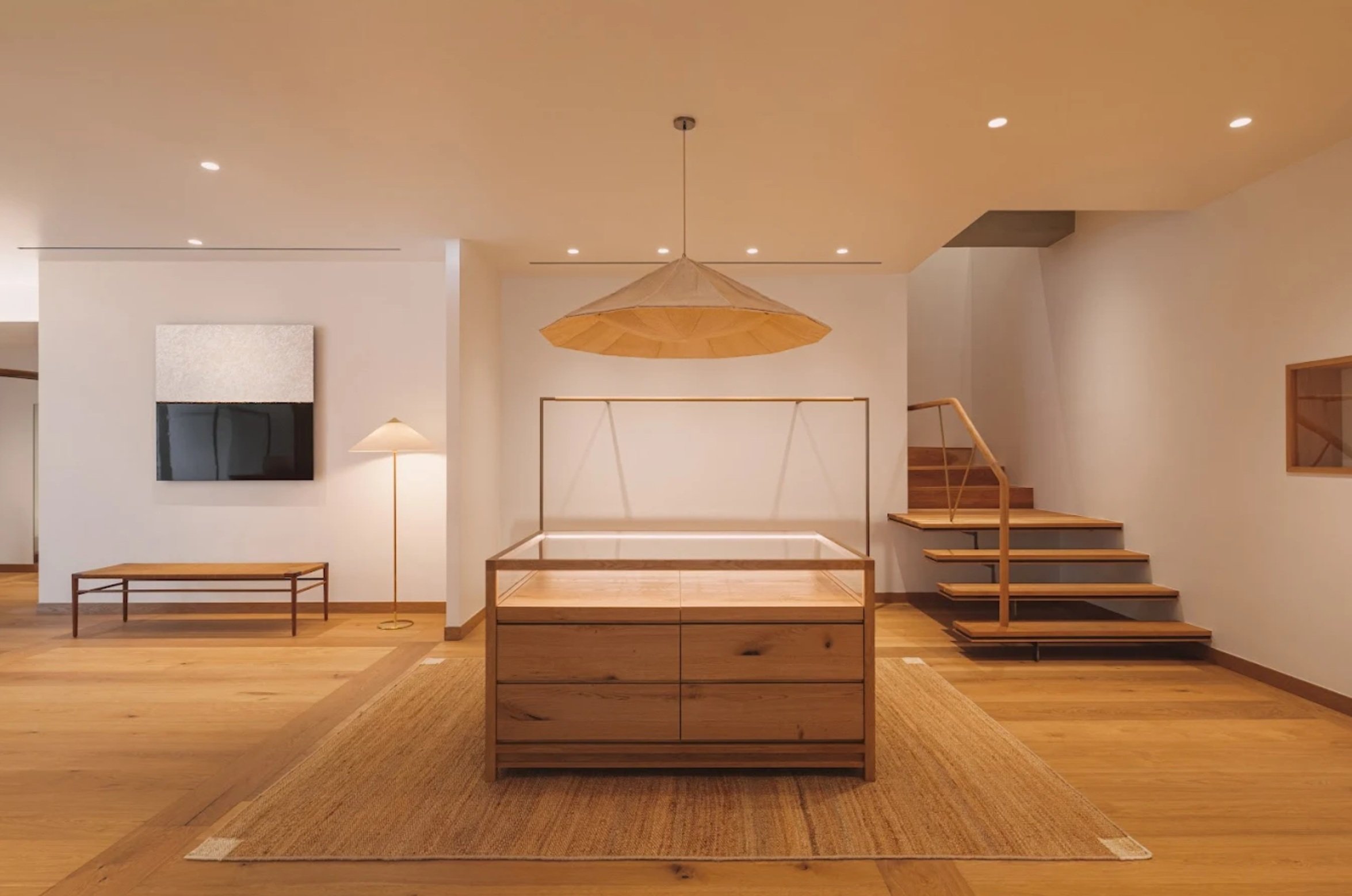 A minimalist interior space featuring a wooden display table, a wooden bench, a standing lamp, a large pendant light, a staircase with wooden steps, and a wall-mounted television, all set on a wooden floor with a textured rug.