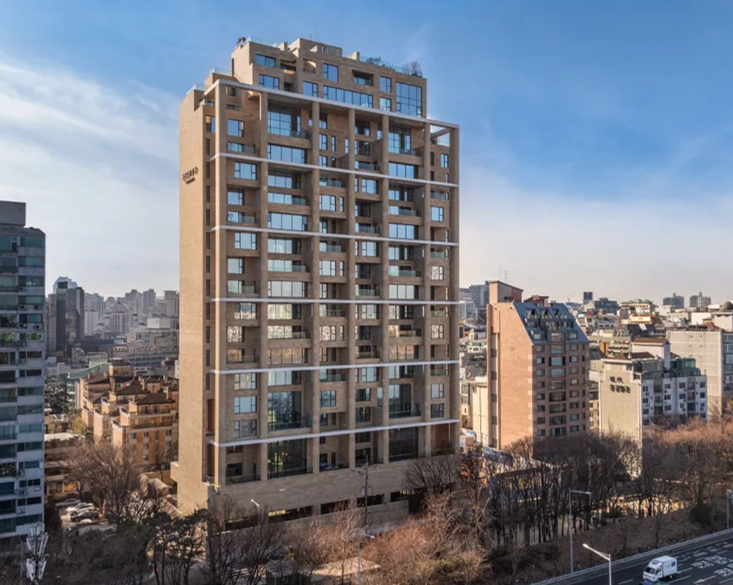 Tall modern apartment building in a city with a blue sky, buildings in the background, and some trees and cars below.