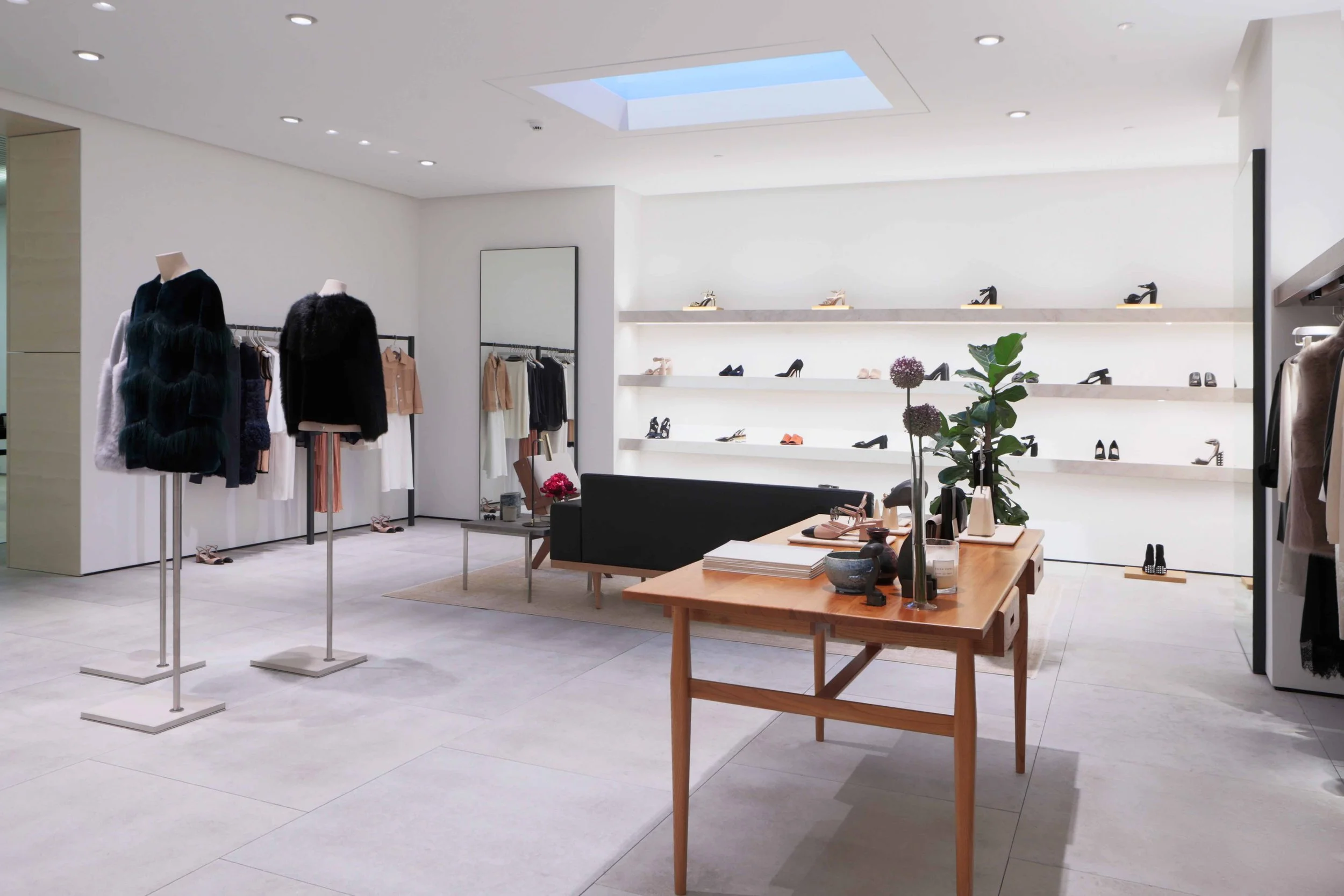 Interior view of a shoe store with shelves displaying high heels and sandals, display tables with accessories, and clothing racks with jackets and skirts, all in a bright, modern space.