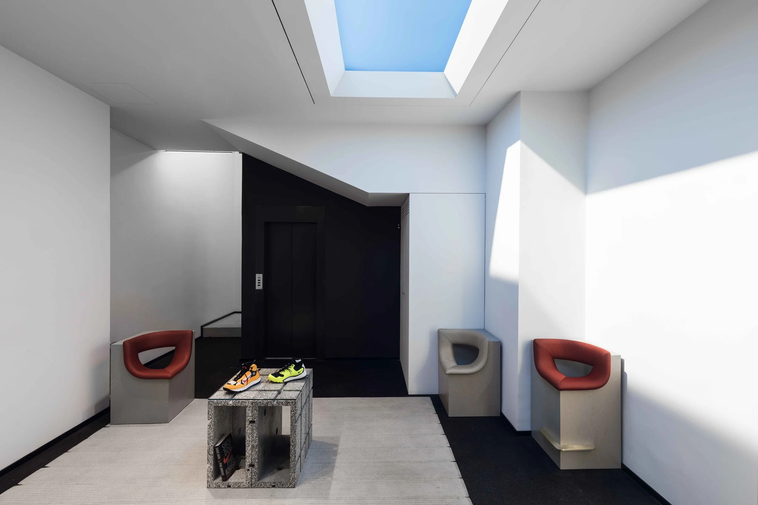 Modern interior space with white walls and a skylight ceiling, featuring red and beige chairs, a granite table with shoes, and a black elevator door.