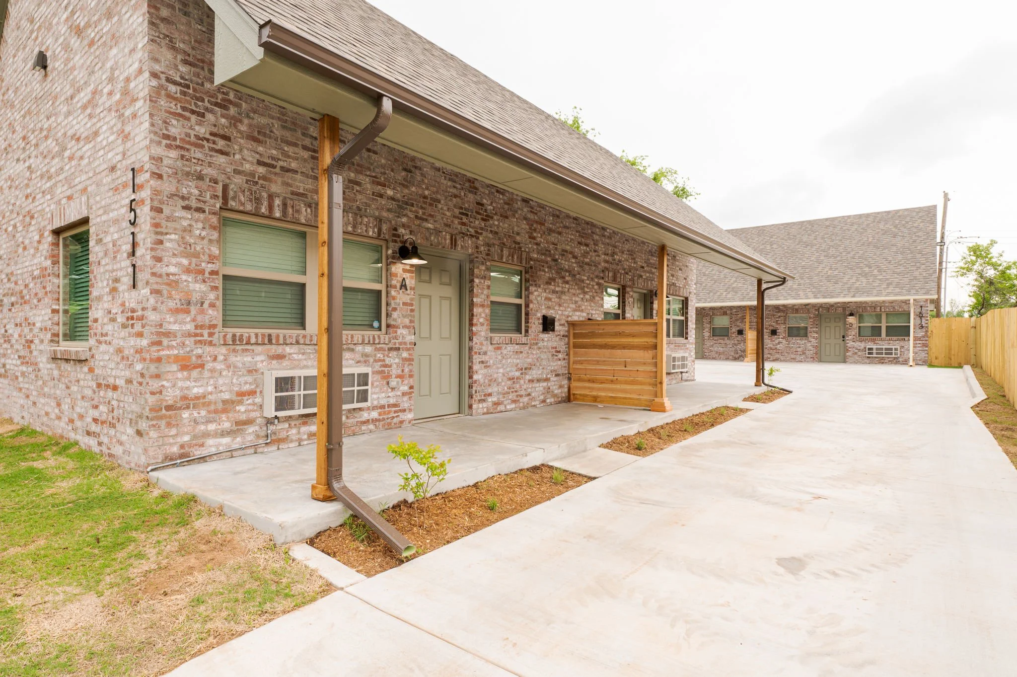 A row of brick houses with small front porches, green doors, and windows, with a concrete sidewalk and small patches of grass and garden beds in front. There is a brown downspout attached to a wooden post, and a black outdoor light fixture next to the door.