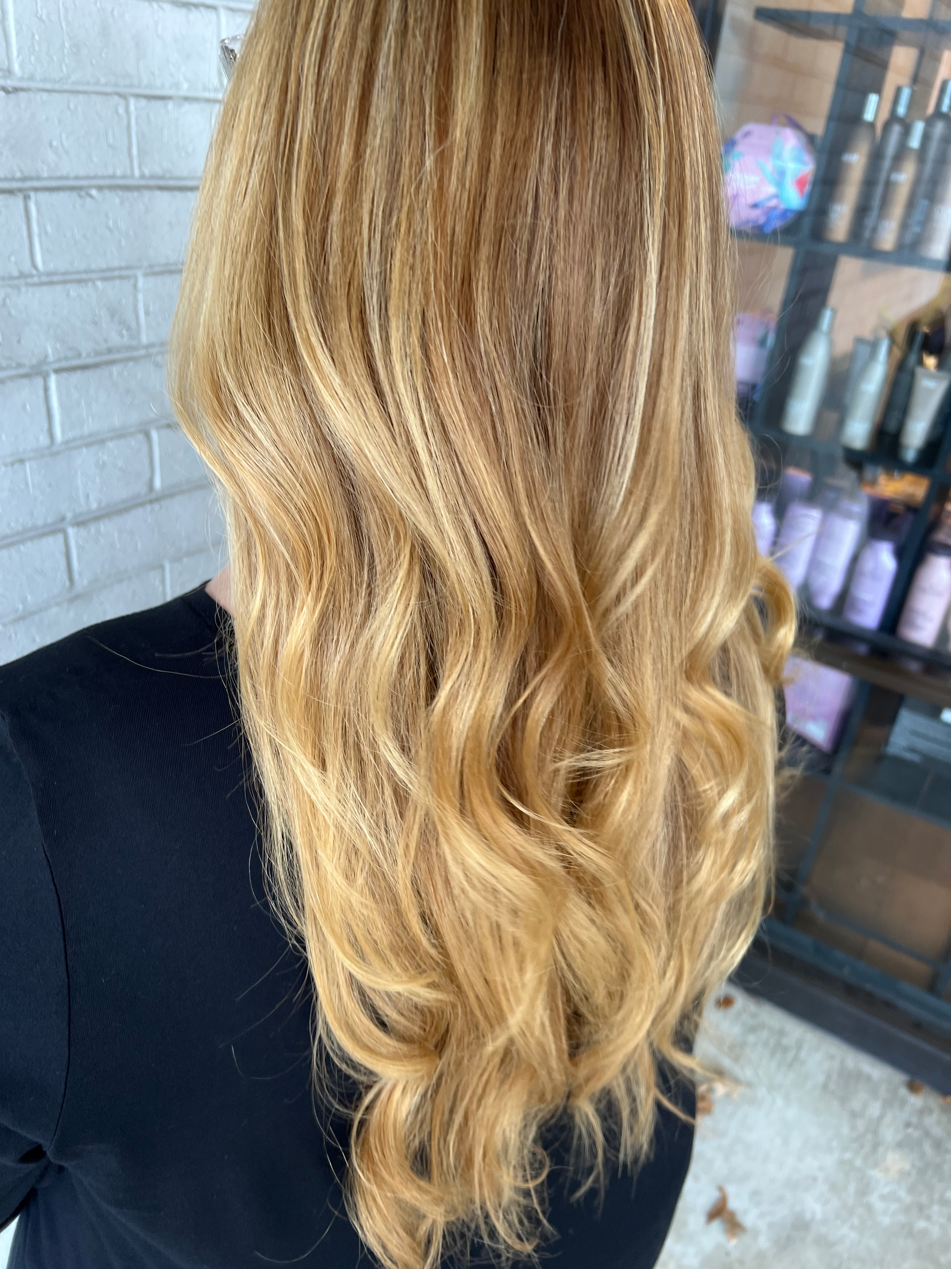 Wavy blonde hair cascading down a person's back, with a background featuring hair products on shelves.