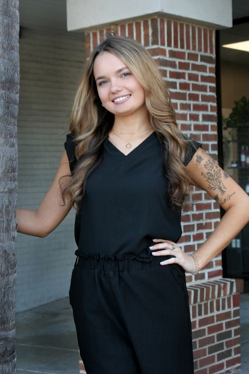 Person standing outdoors smiling, wearing a black blouse and pants, with a tattoo on the arm, and a hand resting on a brick wall.