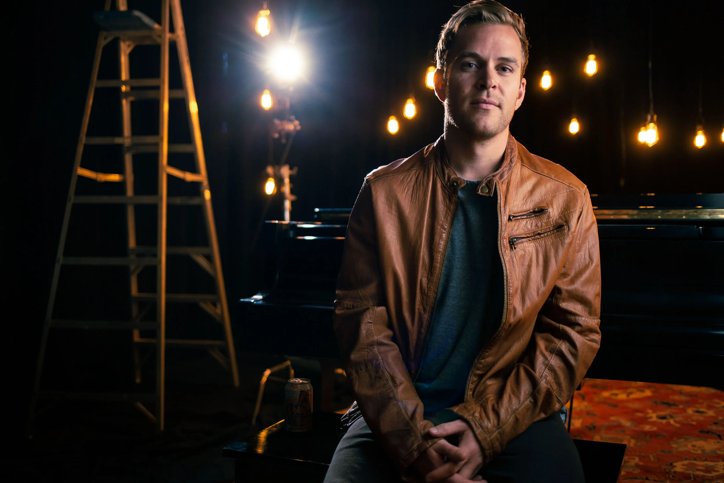 Actor on a lit stage beside a wooden ladder and Edison bulbs