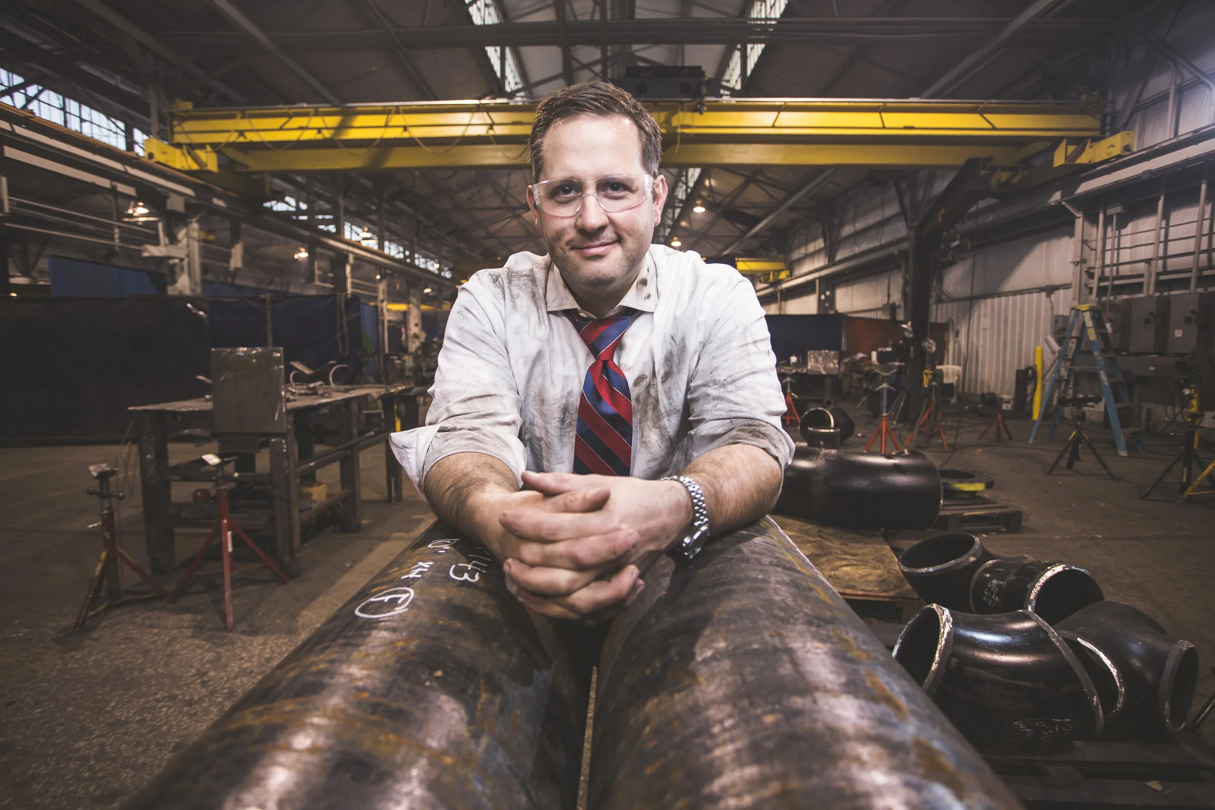 Man in shirt and tie leaning on steel pipe in a fabrication shop