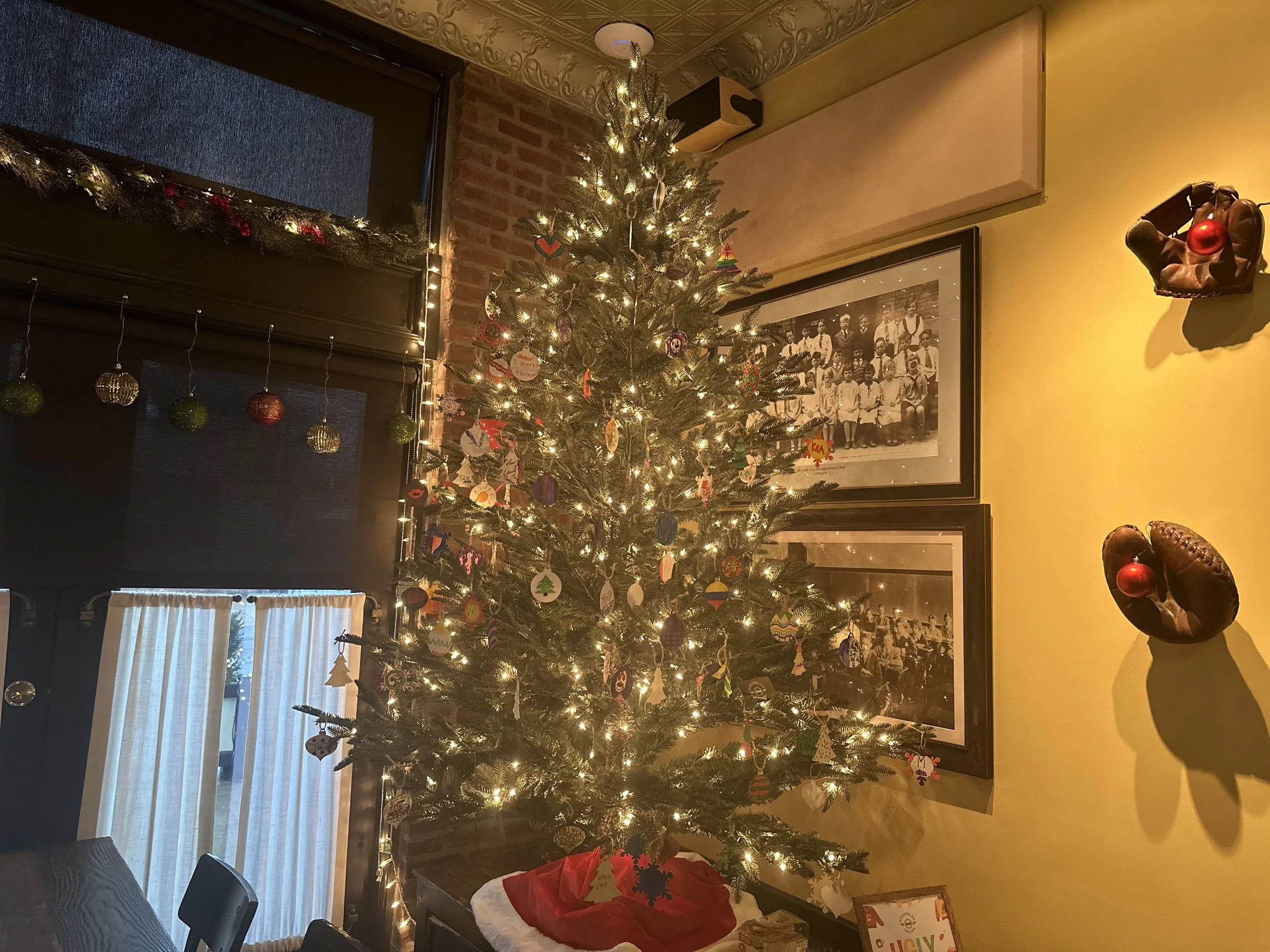 Living room with decorated Christmas tree illuminated with lights, surrounded by holiday decorations, framed black-and-white photographs on yellow wall, and decorative leather baseball gloves with Christmas ornaments on the wall.