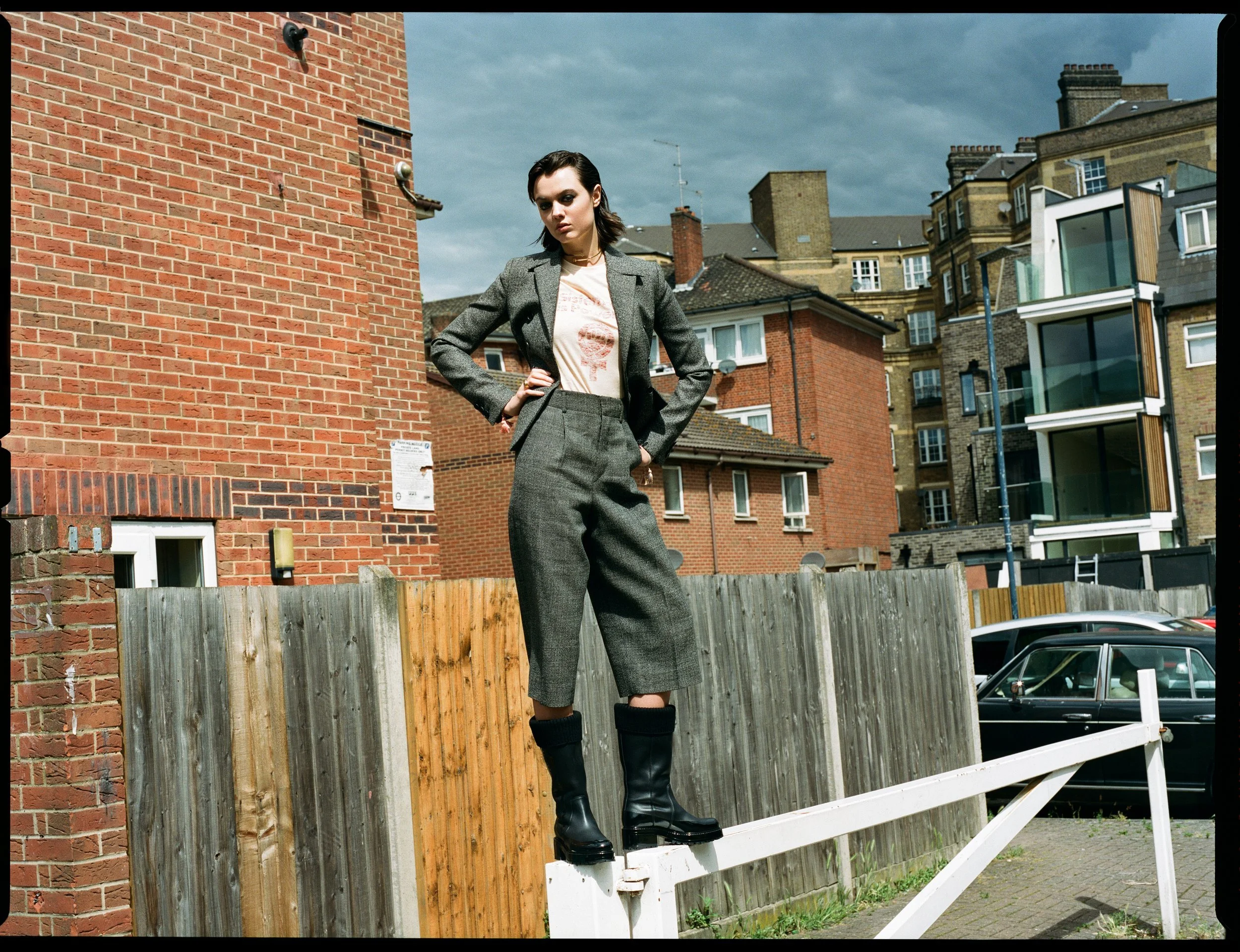 A woman in a gray suit wearing black boots standing on a white barrier in an urban area with brick and modern buildings in the background.