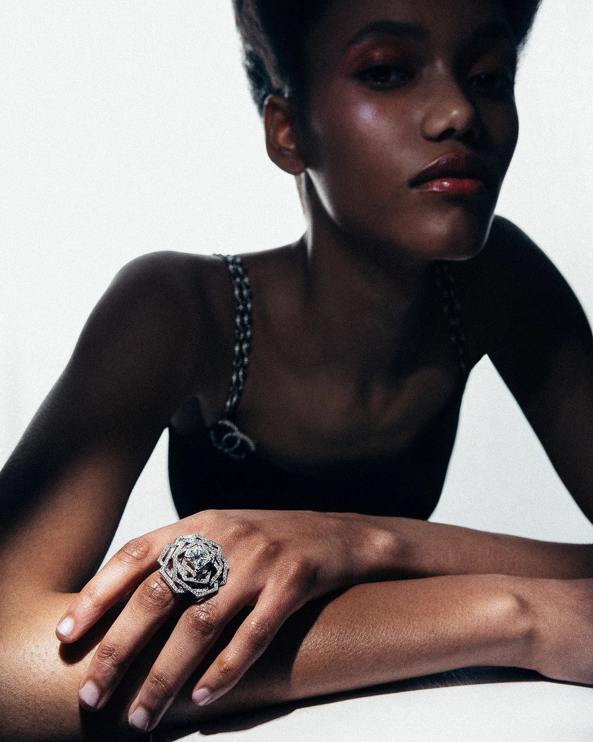 A woman with dark skin and dark hair wearing a black top with chain straps, resting her arms on a surface, showcasing a large silver ring shaped like a rose on her finger.