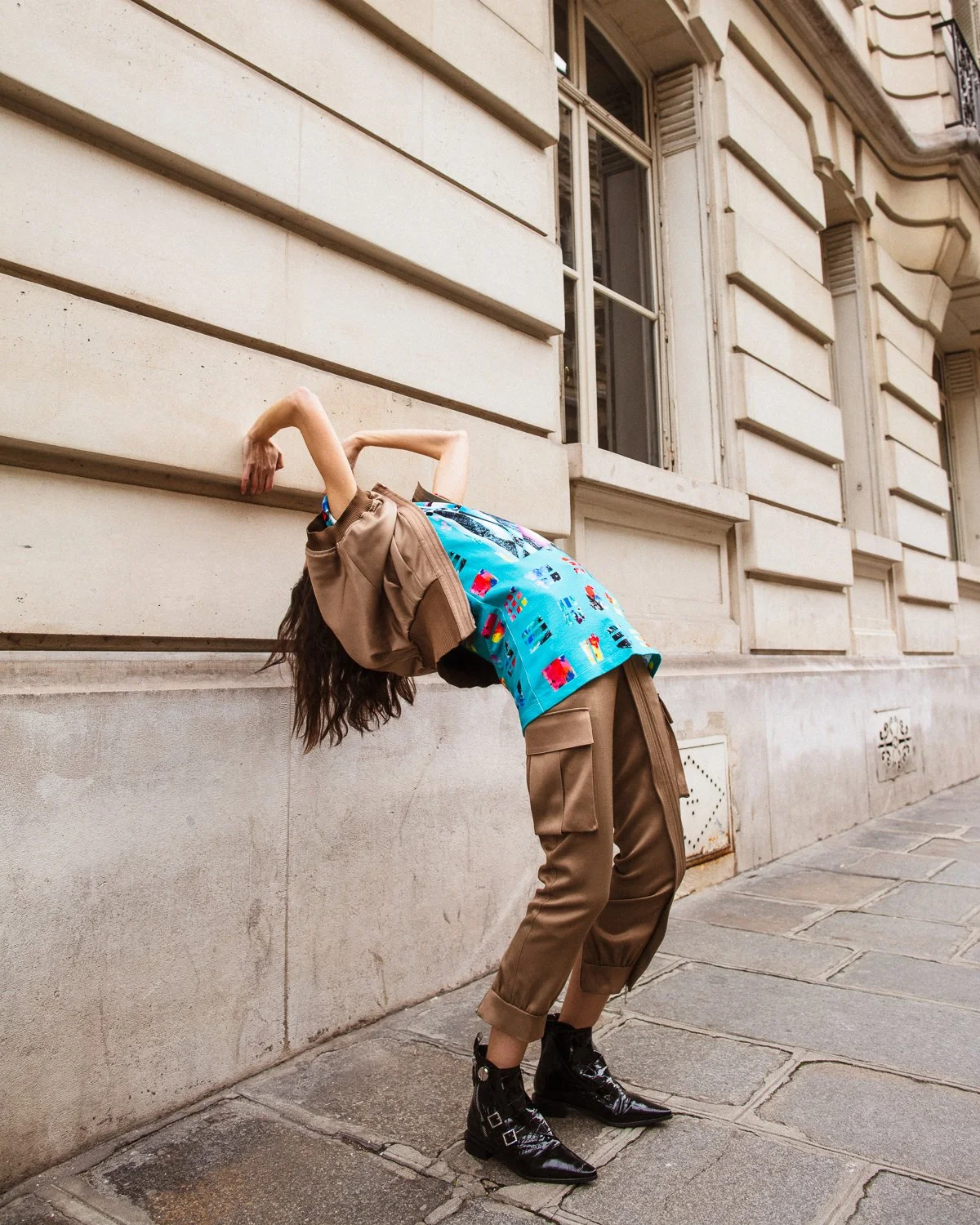 Person bending backwards against a stone wall on a city sidewalk, wearing a colorful blue shirt, brown pants, and black boots, with disheveled hair and a brown jacket draped over their shoulders.