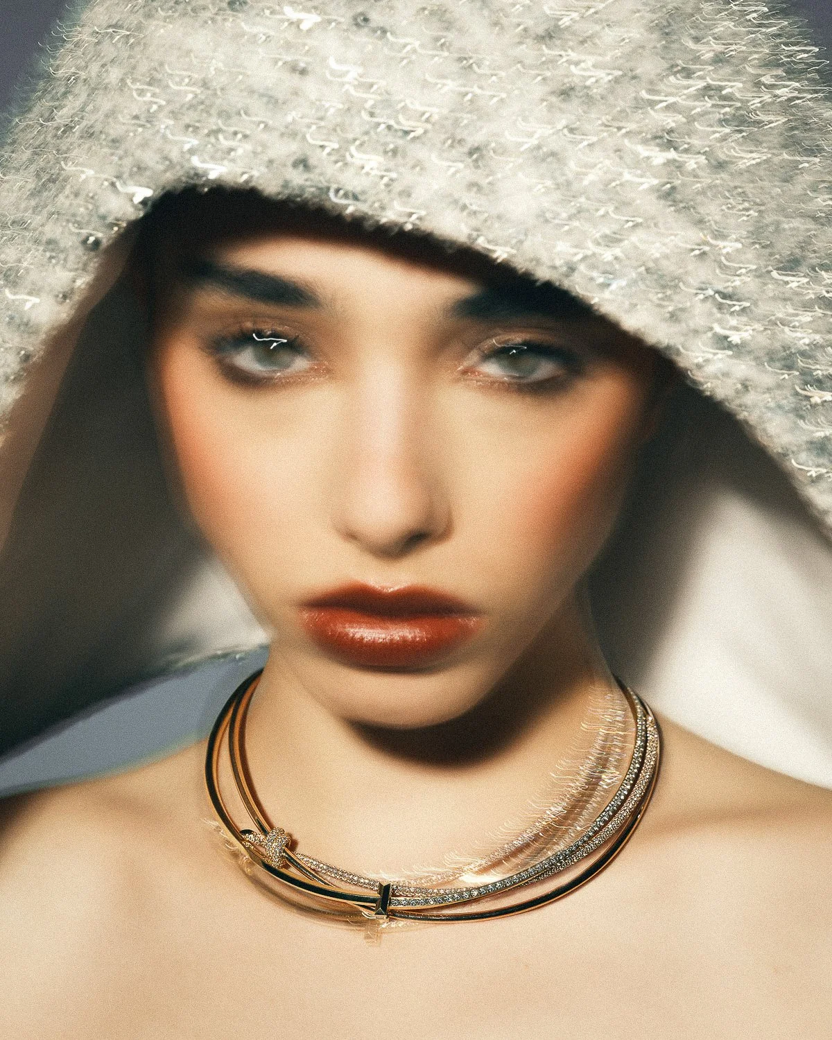 Close-up photo of a woman with makeup, wearing a sparkly gray hat and layered necklaces, looking into the camera.