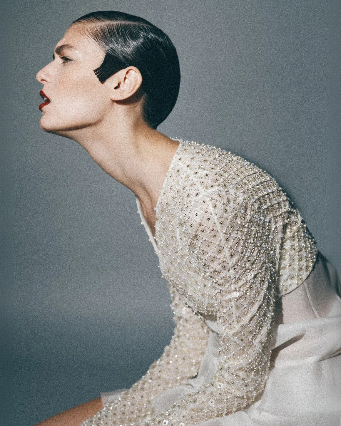 A woman with dark, sleek, side-parted hair, wearing red lipstick and a beaded dress with embroidery, posing against a neutral background.